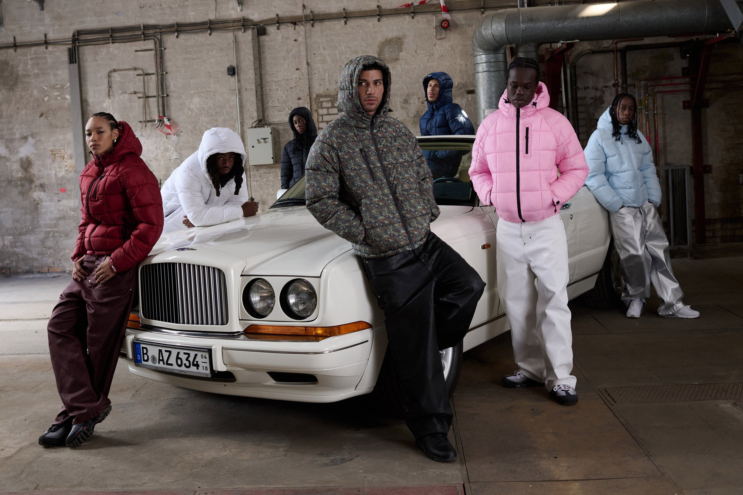 Six people wearing colorful puffer jackets and baggy pants pose around a white vintage car in an industrial garage with concrete walls and exposed pipes.