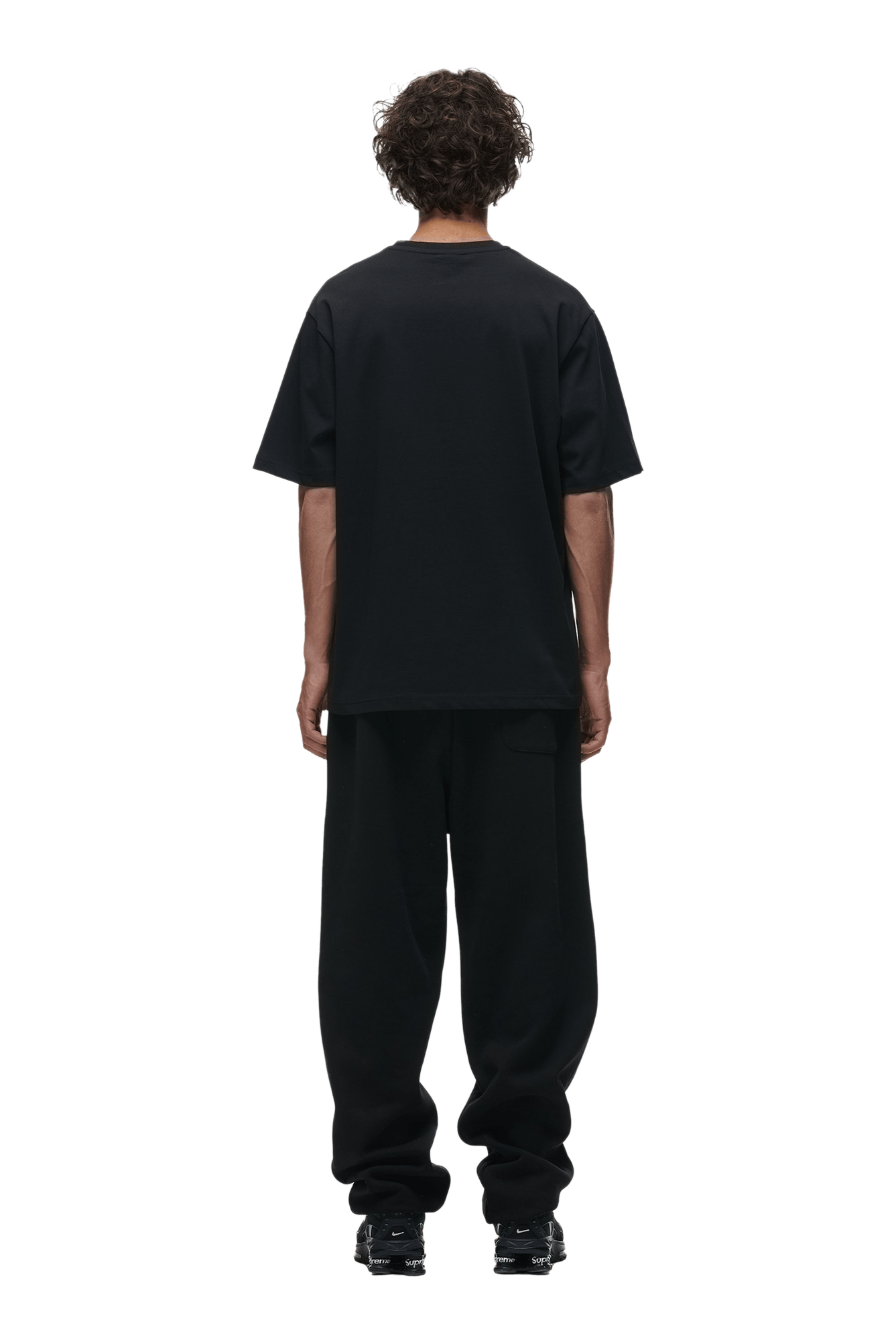 A person with curly hair stands facing away, wearing the 6PM T-SHIRT BLACK, paired with black pants and shoes, on a transparent background.