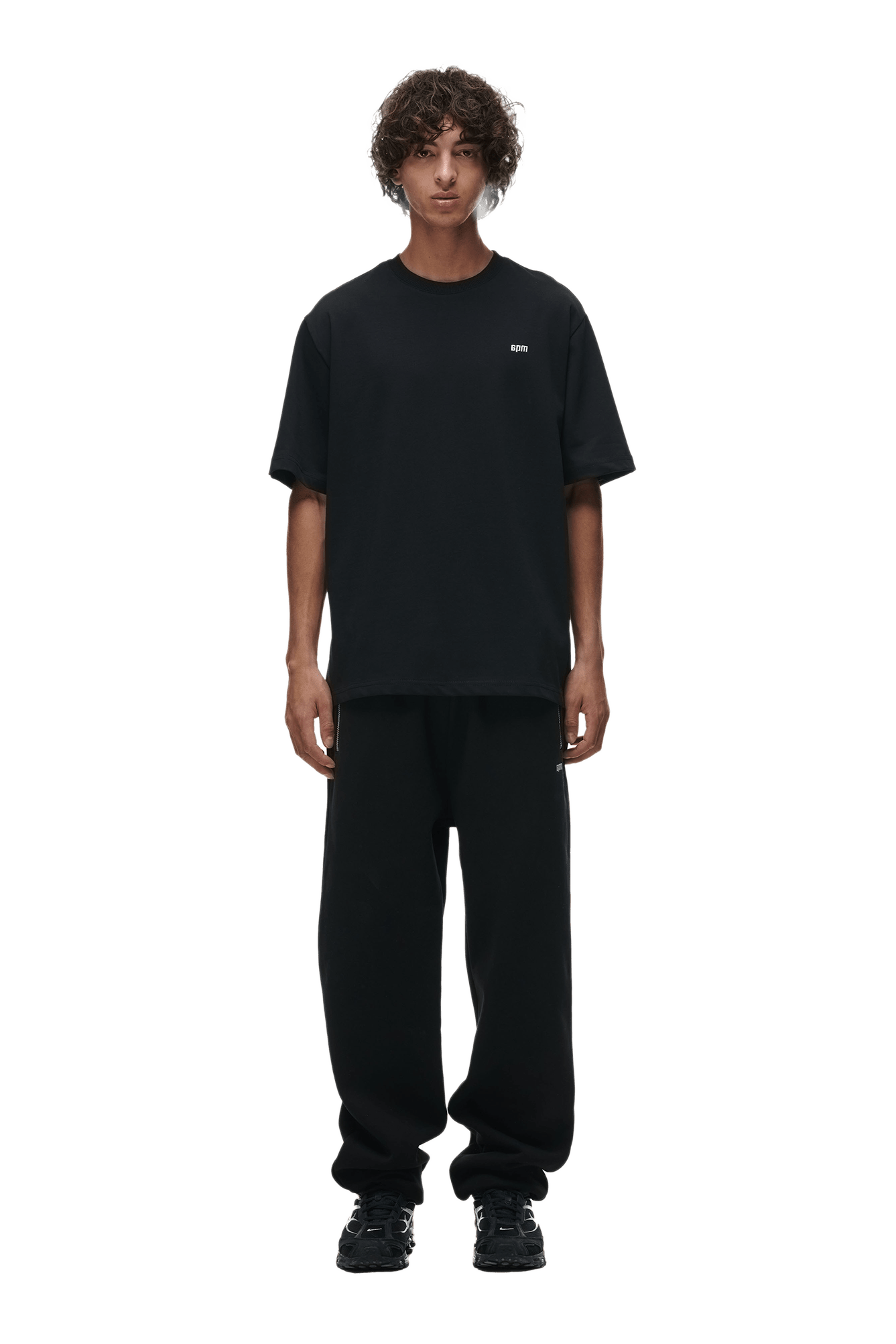 A person with curly hair faces forward, wearing the 6PM T-SHIRT BLACK, black sweatpants, and black sneakers against a plain background.