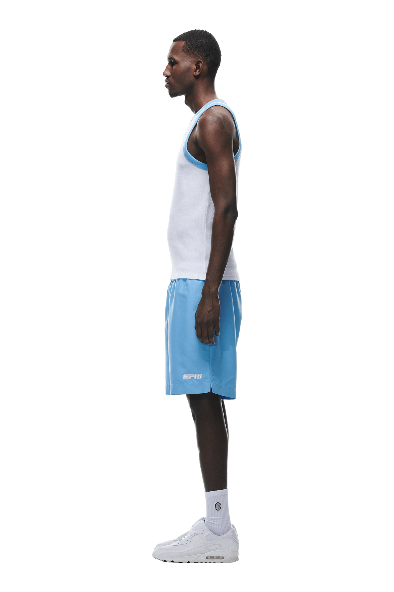 A man stands in profile against a white background, wearing the 6PM WAFFLE KNIT TANKTOP WHITE, light blue athletic shorts, white socks, and white sneakers.