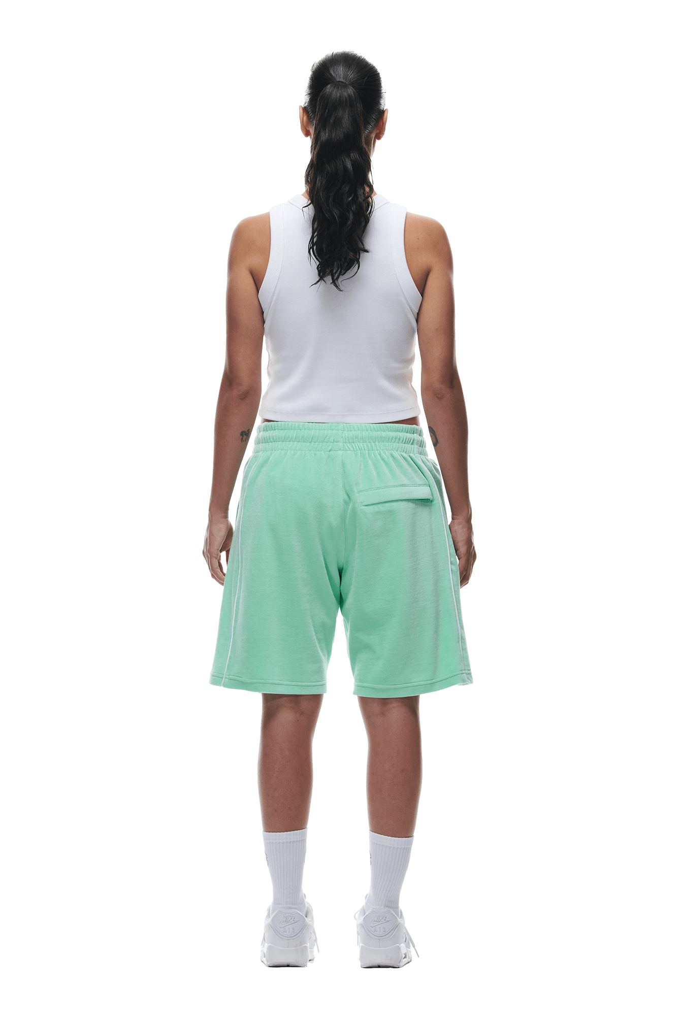 A person with long dark hair in a ponytail is facing away, wearing a white tank top, 6PM VELOUR SHORTS LIGHT MOSS, white socks, and white sneakers against a plain white background.