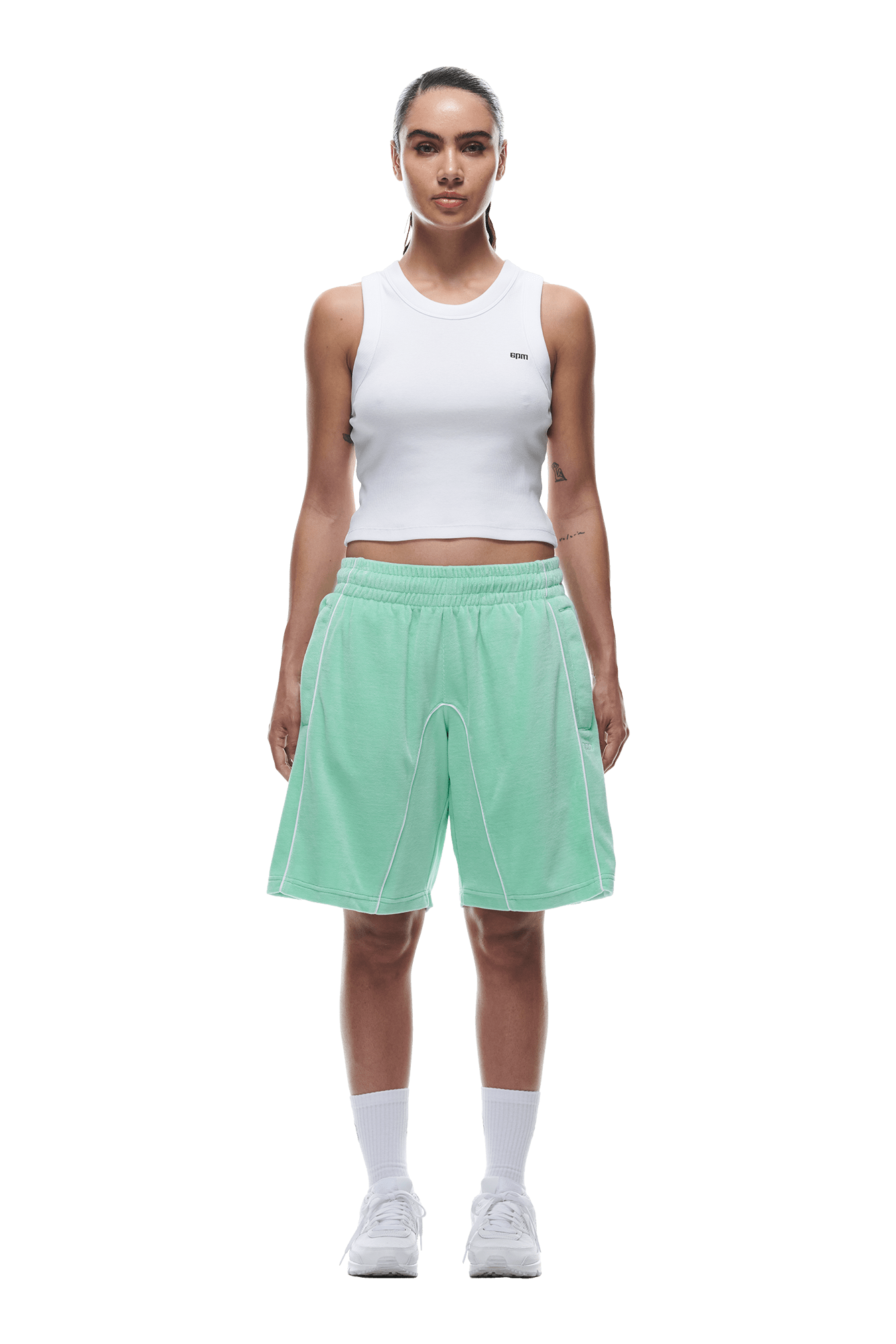 A woman faces forward, wearing a white sleeveless crop top, 6PM VELOUR SHORTS LIGHT MOSS, white socks, and white sneakers against a plain white background.
