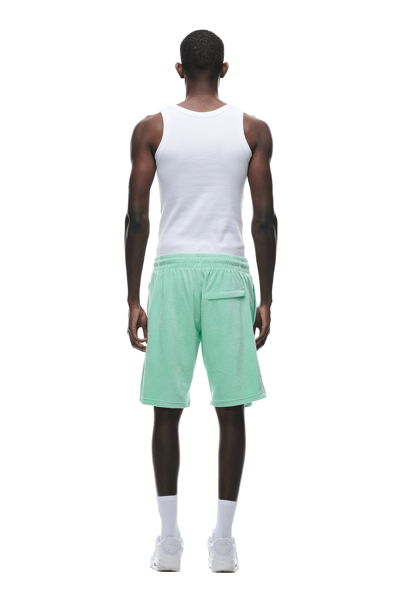 A man stands facing away, wearing a white tank top, 6PM VELOUR SHORTS LIGHT MOSS, white socks, and white sneakers against a plain white background.