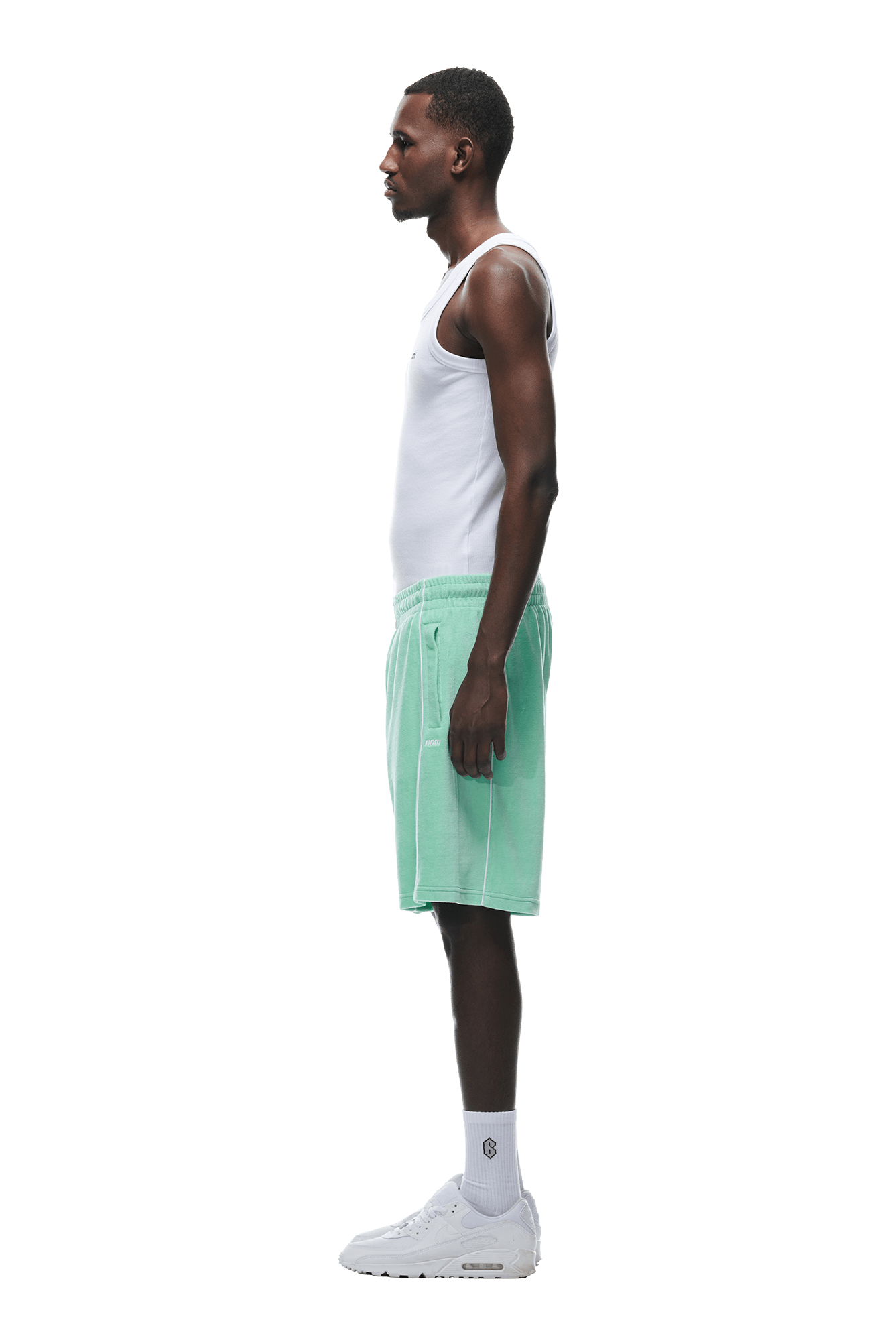 A man stands in profile against a white background, wearing a white tank top, 6PM VELOUR SHORTS LIGHT MOSS, white socks, and white sneakers.
