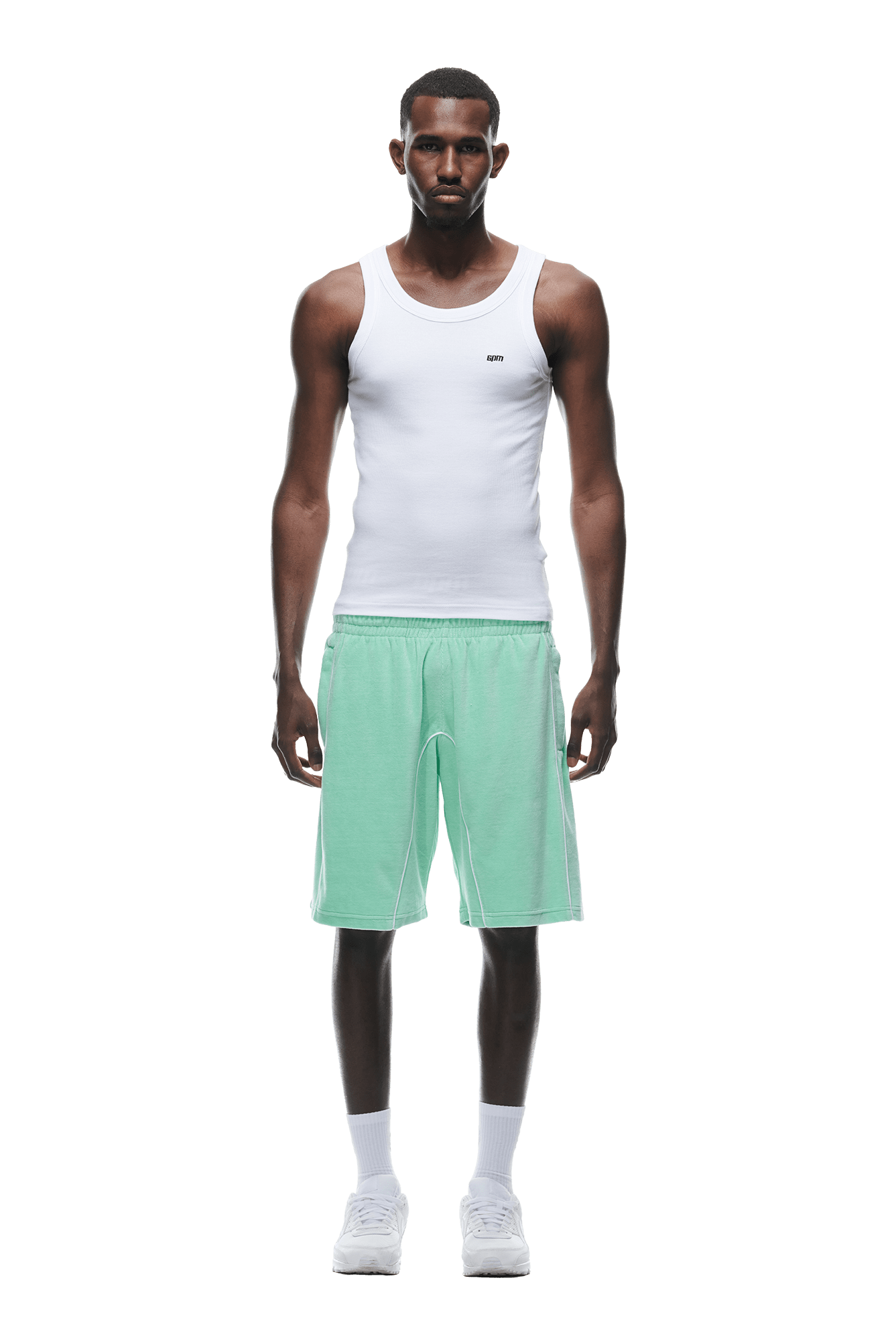 A man faces forward wearing a fitted white tank top, 6PM VELOUR SHORTS LIGHT MOSS, white crew socks, and white sneakers against a plain white background.