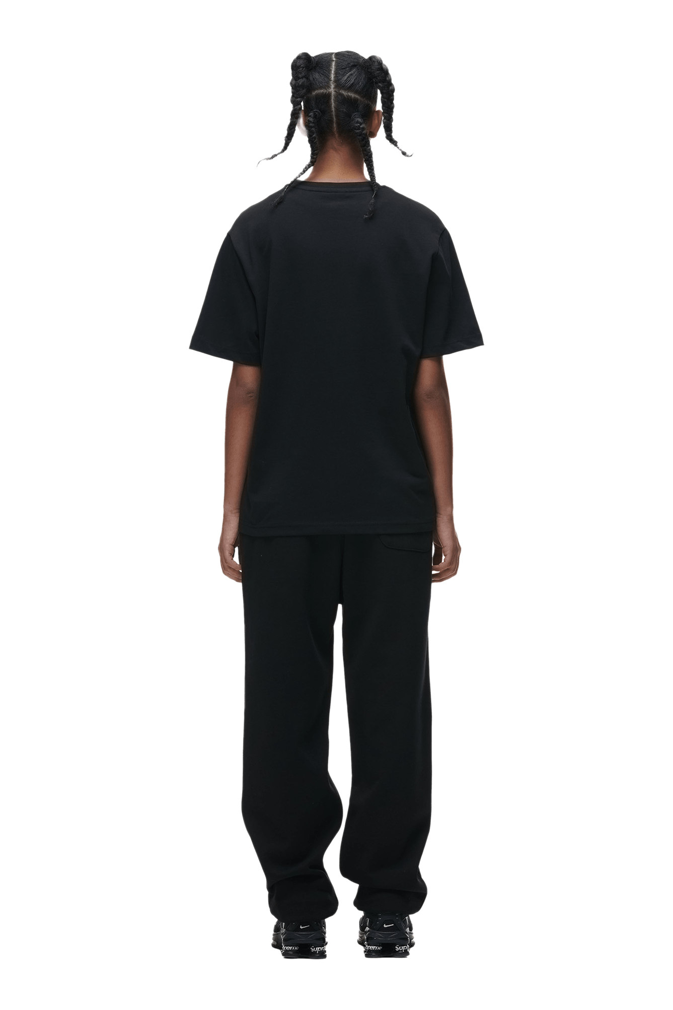 A person with braided hair faces away, wearing the 6PM T-SHIRT BLACK paired with black pants and shoes, set against a plain green background.