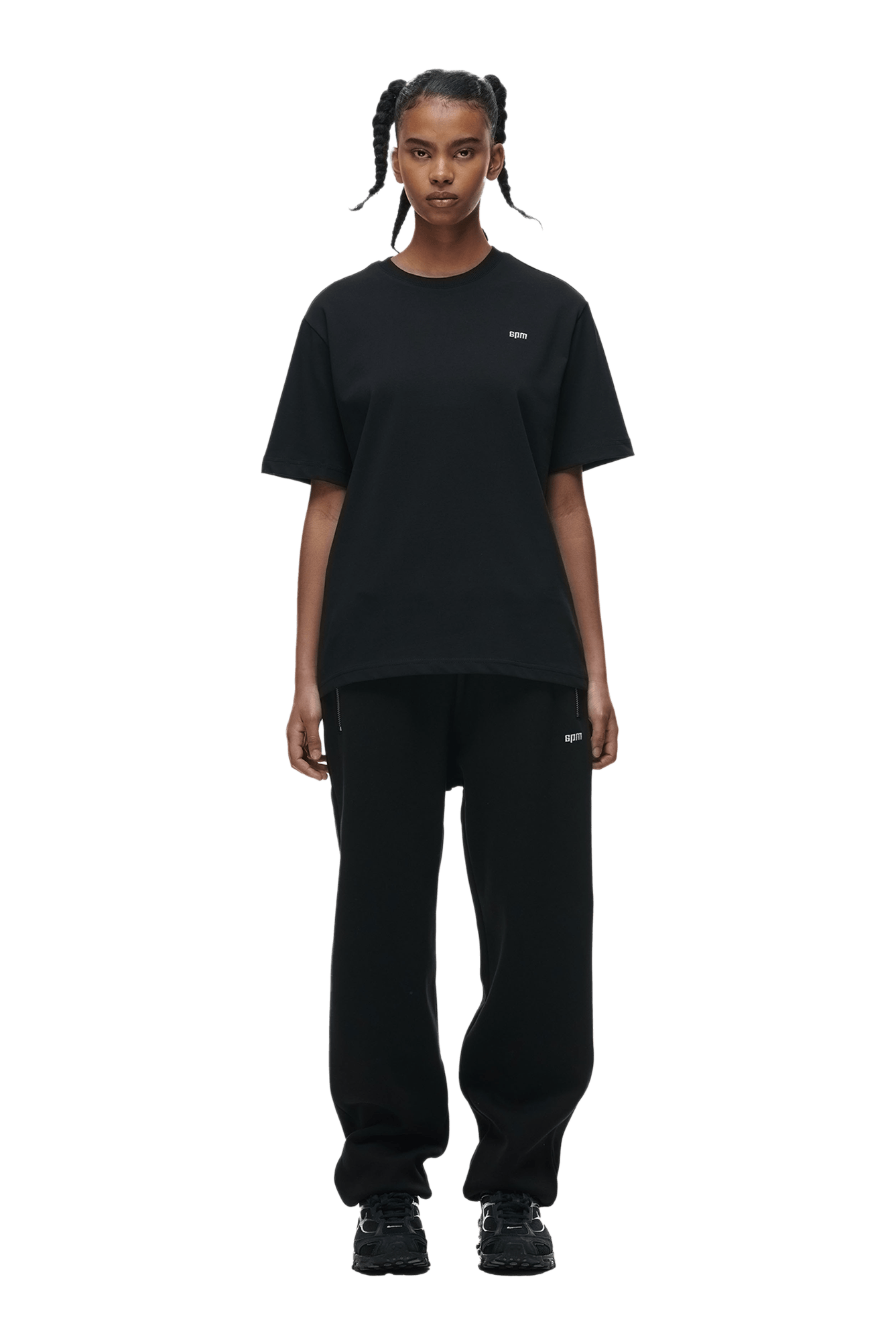 A person with braided hair stands facing forward, wearing a 6PM T-SHIRT BLACK, black sweatpants, and black sneakers against a plain green background.