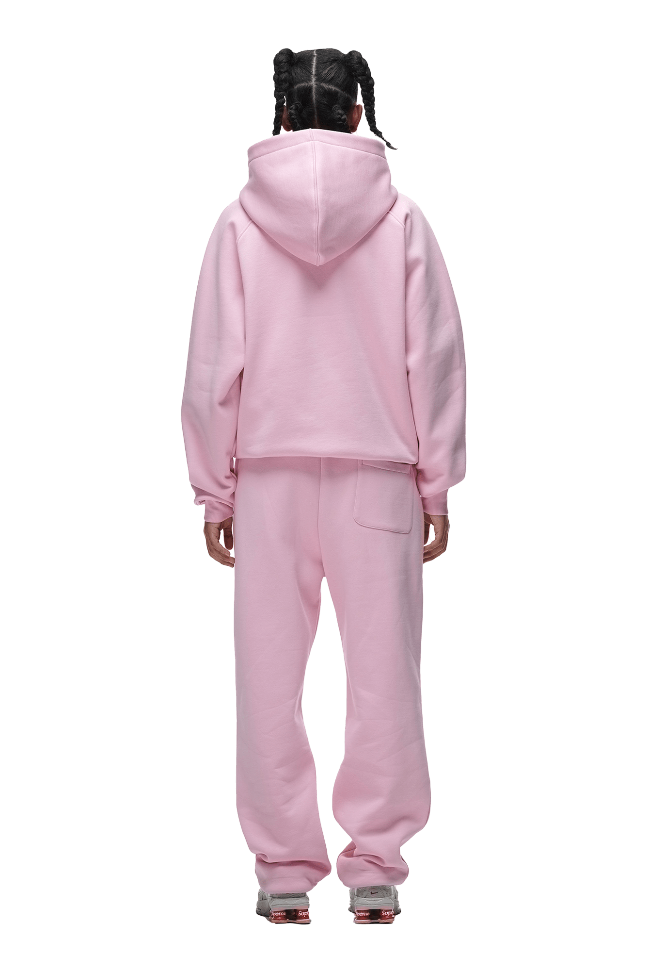A person with braided hair, seen from behind, wears the 6PM HOODIE BABY PINK—an oversized raglan-sleeved hoodie with a kangaroo pocket, matched with pink sweatpants and white sneakers. The transparent background highlights this everyday outfit.