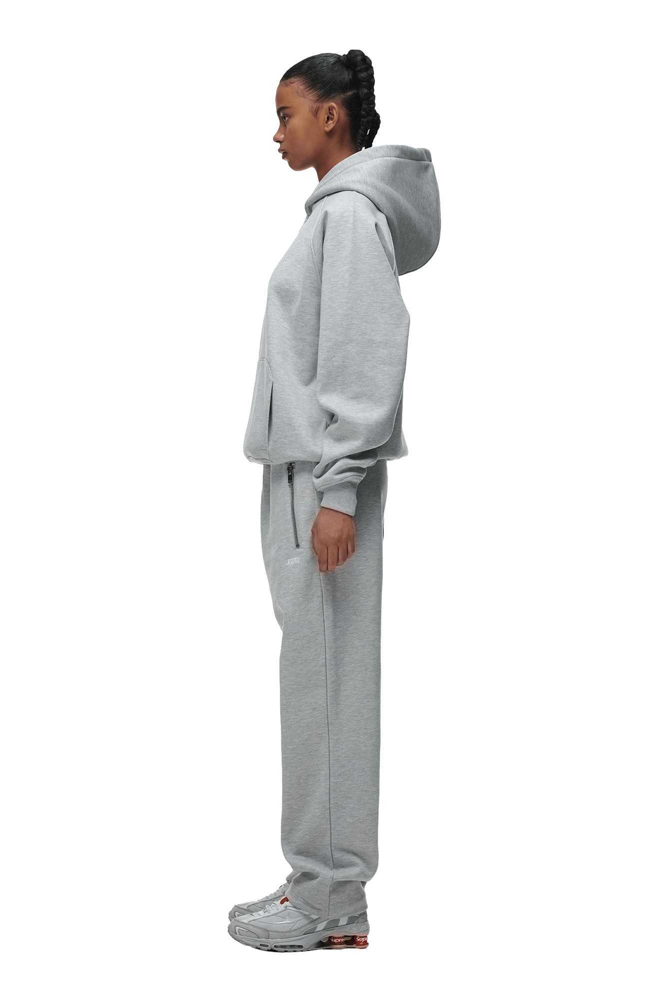 A woman stands in profile wearing the 6PM HOODIE GREY MELANGE with matching sweatpants and gray sneakers, looking straight ahead against a transparent background—ideal for daily wear.