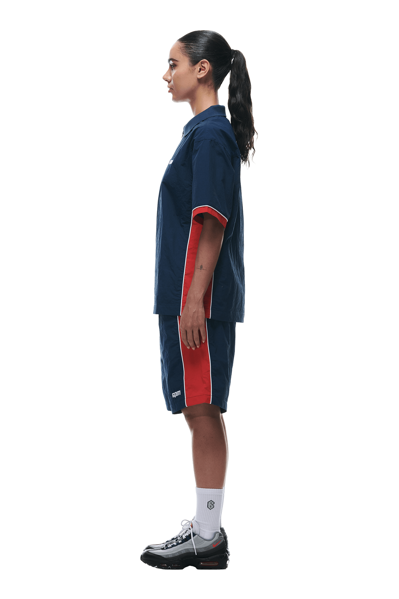 A person stands in profile against a white background, wearing the 6PM WARM UP NYLON HALF ZIP NIGHT SKY matching shirt and shorts with red side stripes, white crew socks, black sneakers, and a long ponytail.