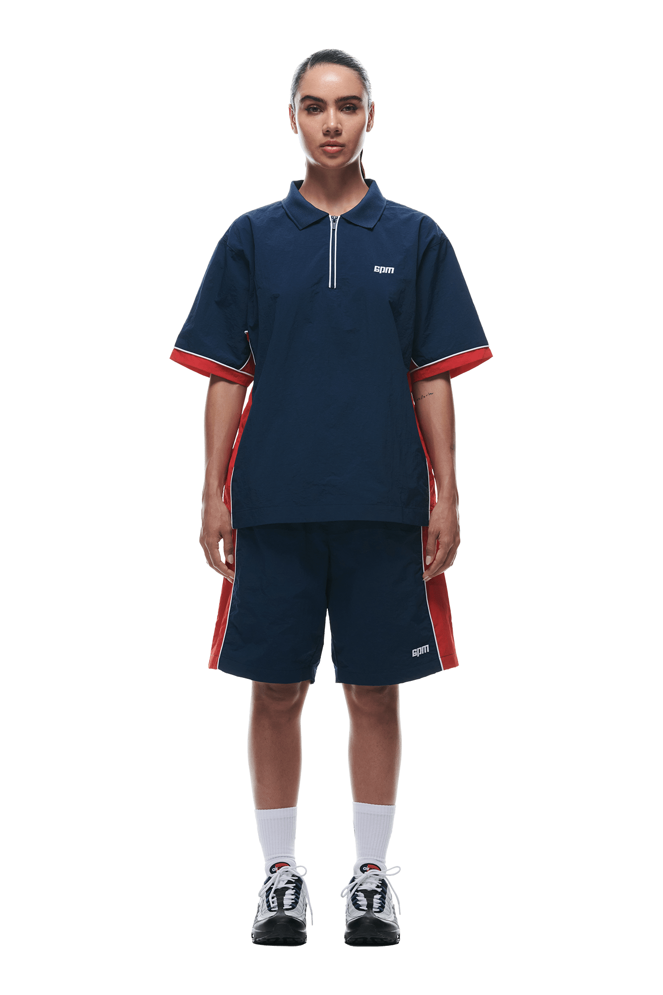 A person stands facing forward in 6PM's WARM UP NYLON HALF ZIP NIGHT SKY shirt and matching shorts with red and white side stripes, white socks, and black sneakers against a plain white background.