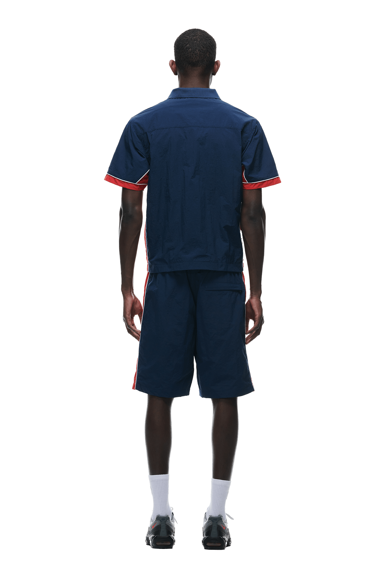 A man stands facing away, wearing the 6PM WARM UP NYLON HALF ZIP NIGHT SKY shirt and matching shorts with red trim, white socks, and black sneakers against a plain white background.