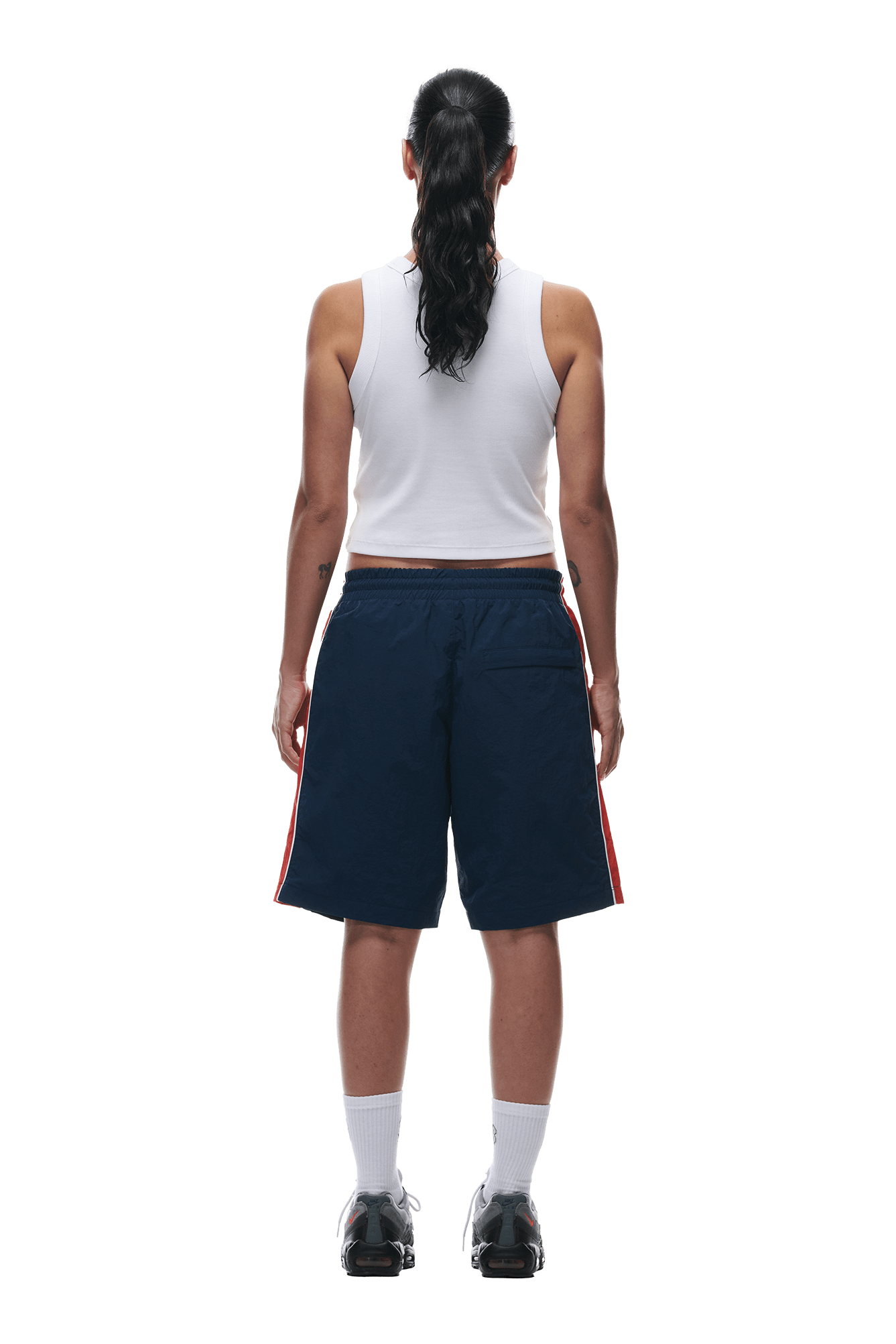 A person with long, dark hair in a ponytail stands facing away, wearing a white sleeveless top, 6PM WARM UP NYLON SHORTS NIGHT SKY, white socks, and black sneakers against a plain white background.