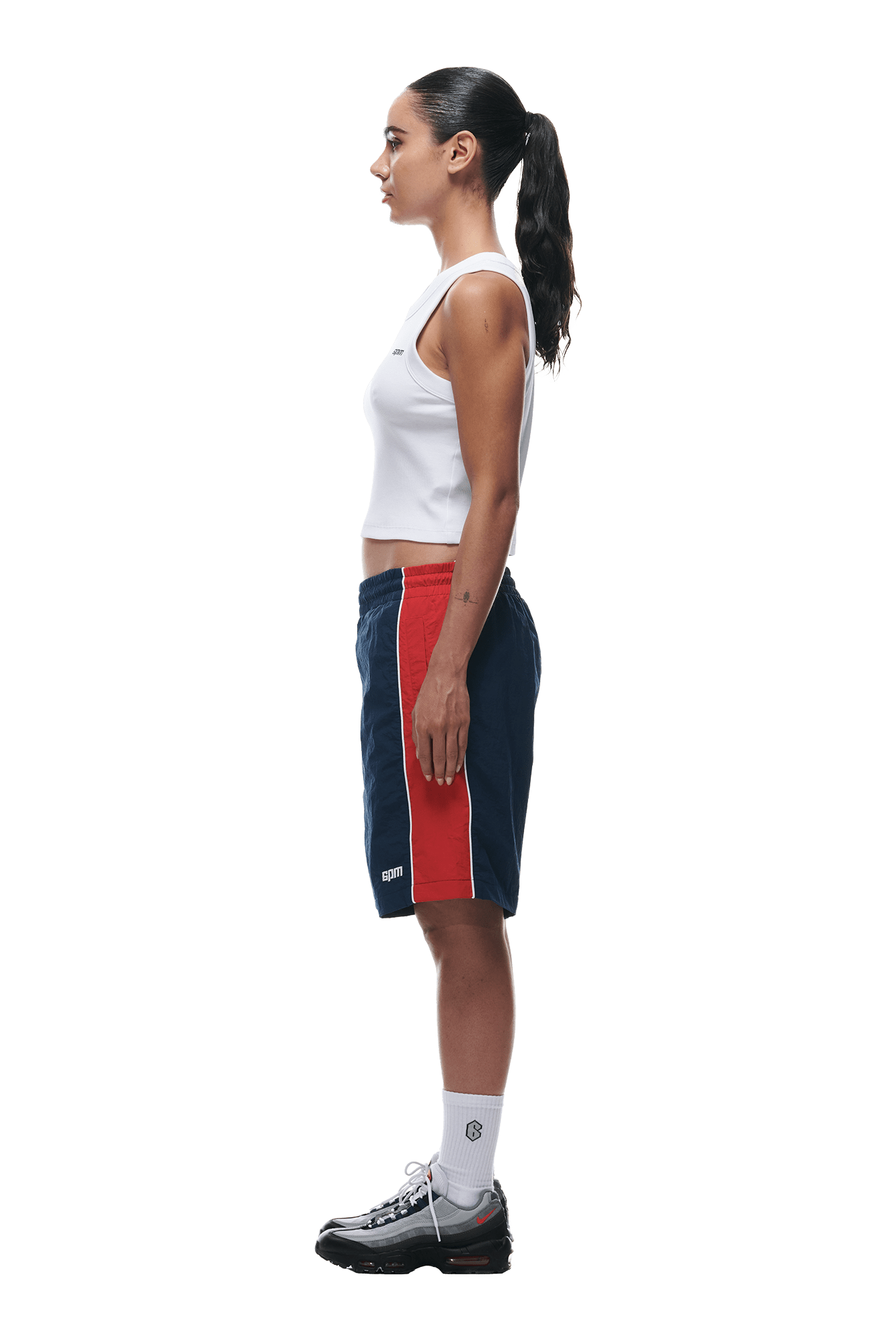 A woman stands in profile against a white background, wearing a white tank top, 6PM WARM UP NYLON SHORTS NIGHT SKY, white socks, and black sneakers. Her hair is tied back in a high ponytail.