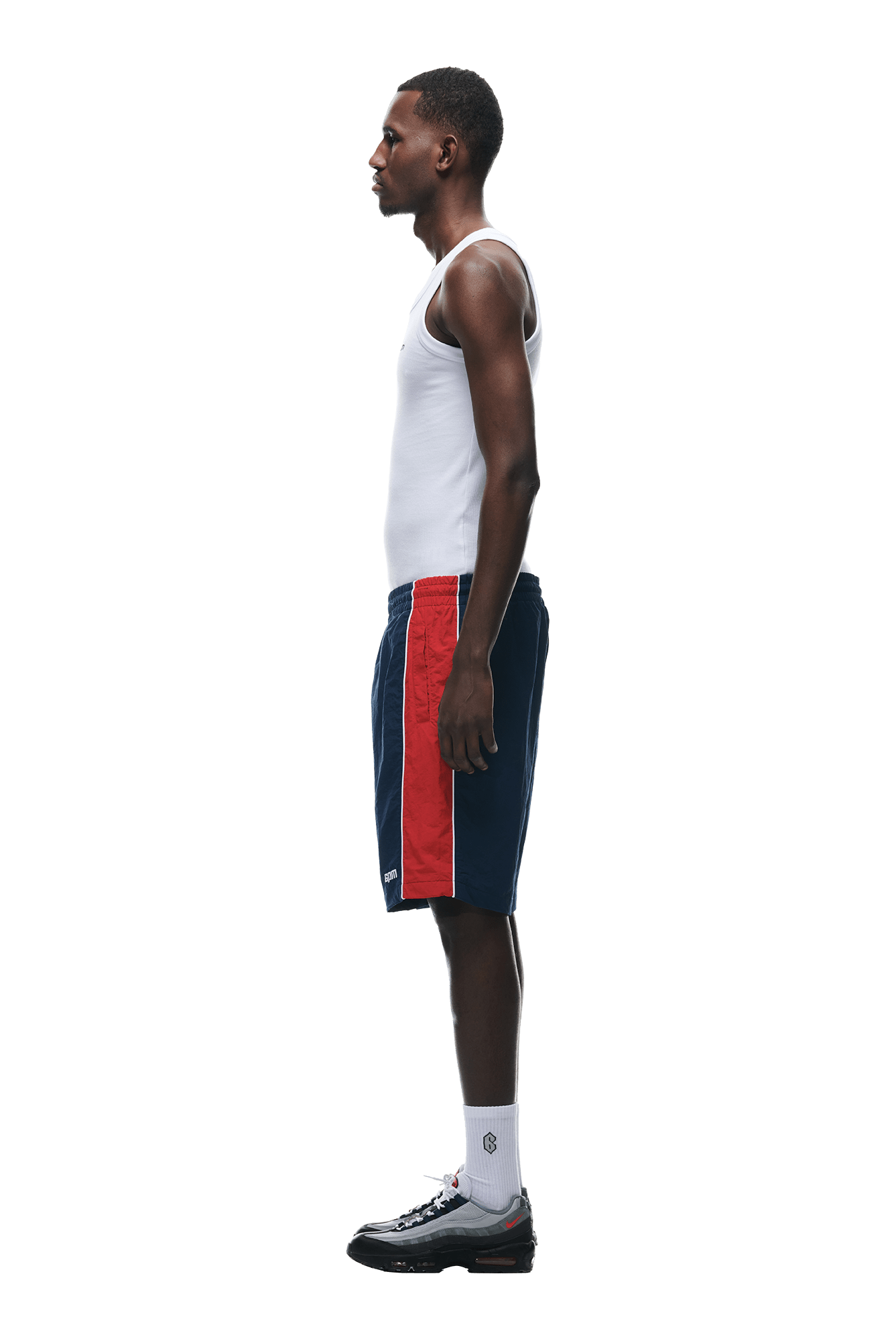 A man stands in profile against a white background, wearing a white tank top, 6PM WARM UP NYLON SHORTS NIGHT SKY, white crew socks, and black athletic shoes.