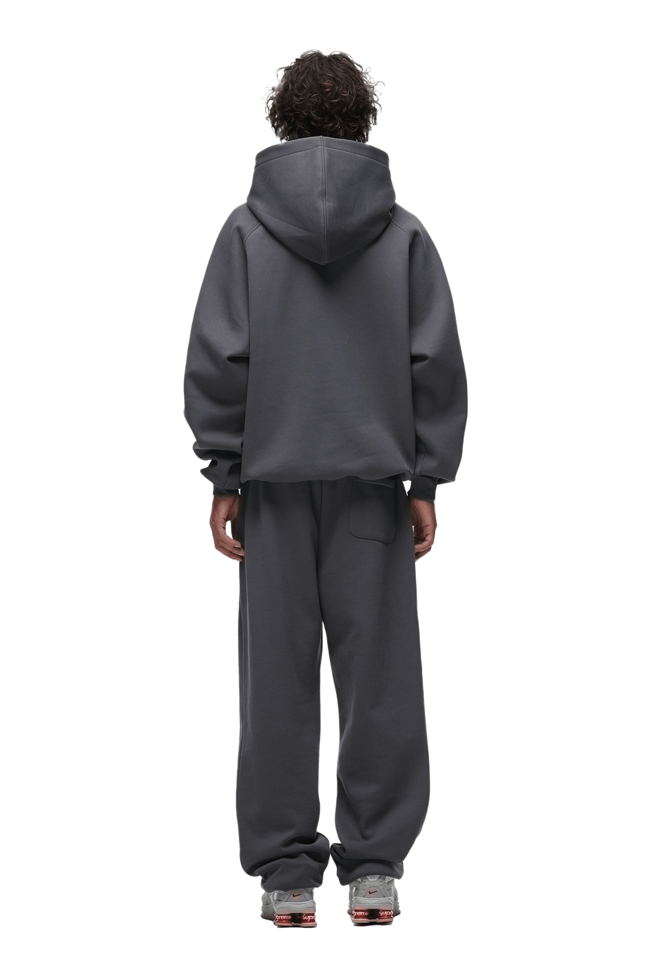 A person with curly hair is facing away, wearing the 6PM HOODIE ANTHRACITE with matching sweatpants and gray sneakers. The background is transparent.