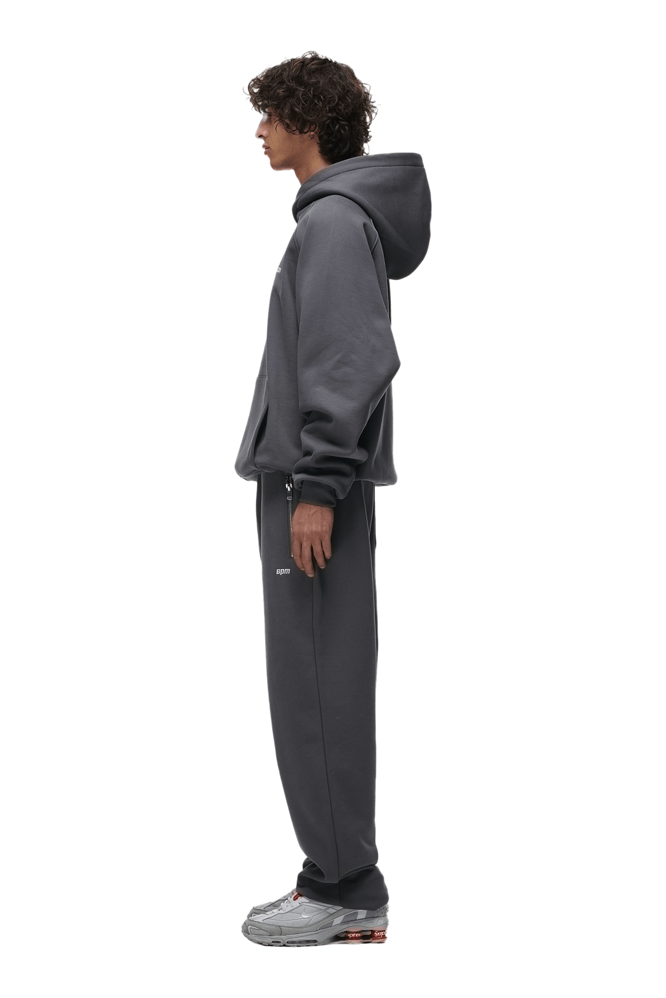 A person with curly hair stands in profile, wearing the 6PM HOODIE ANTHRACITE with Raglan sleeves and a kangaroo pocket, paired with matching sweatpants and gray sneakers—ideal for everyday wear.