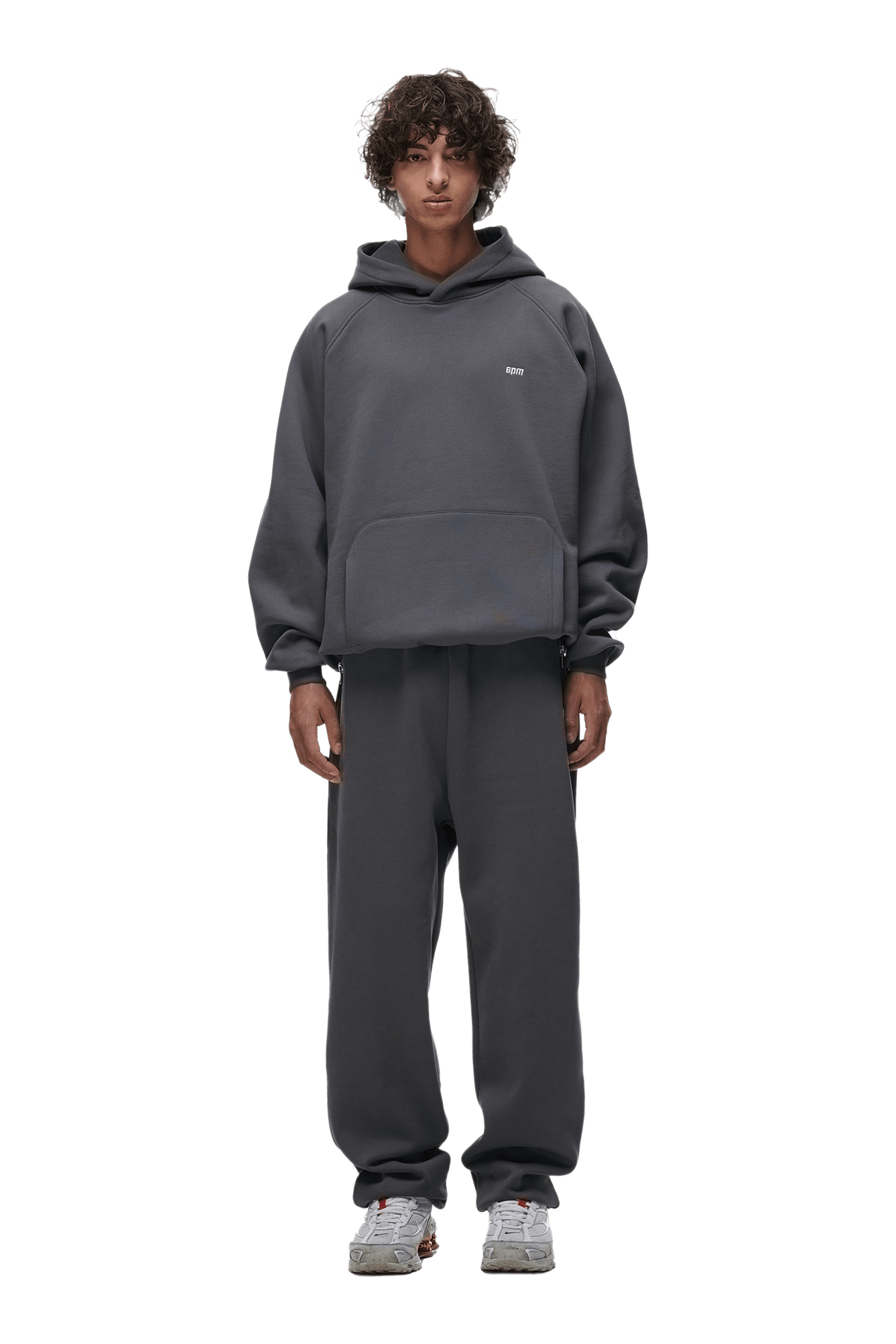 A person with curly hair faces forward wearing the 6PM HOODIE ANTHRACITE— a loose-fit dark gray raglan-sleeved hoodie with kangaroo pocket and matching sweatpants, paired with light gray sneakers. Transparent background, ideal for daily use.