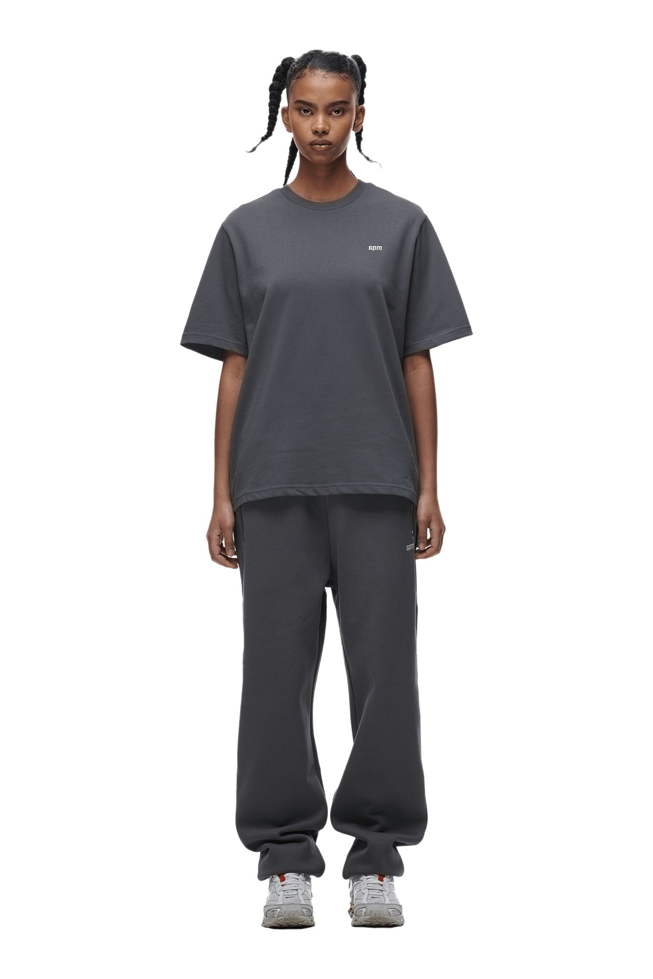 A person with braided hair faces forward, wearing the 6PM T-SHIRT ANTHRACITE paired with matching sweatpants and gray sneakers, set against a plain green background.