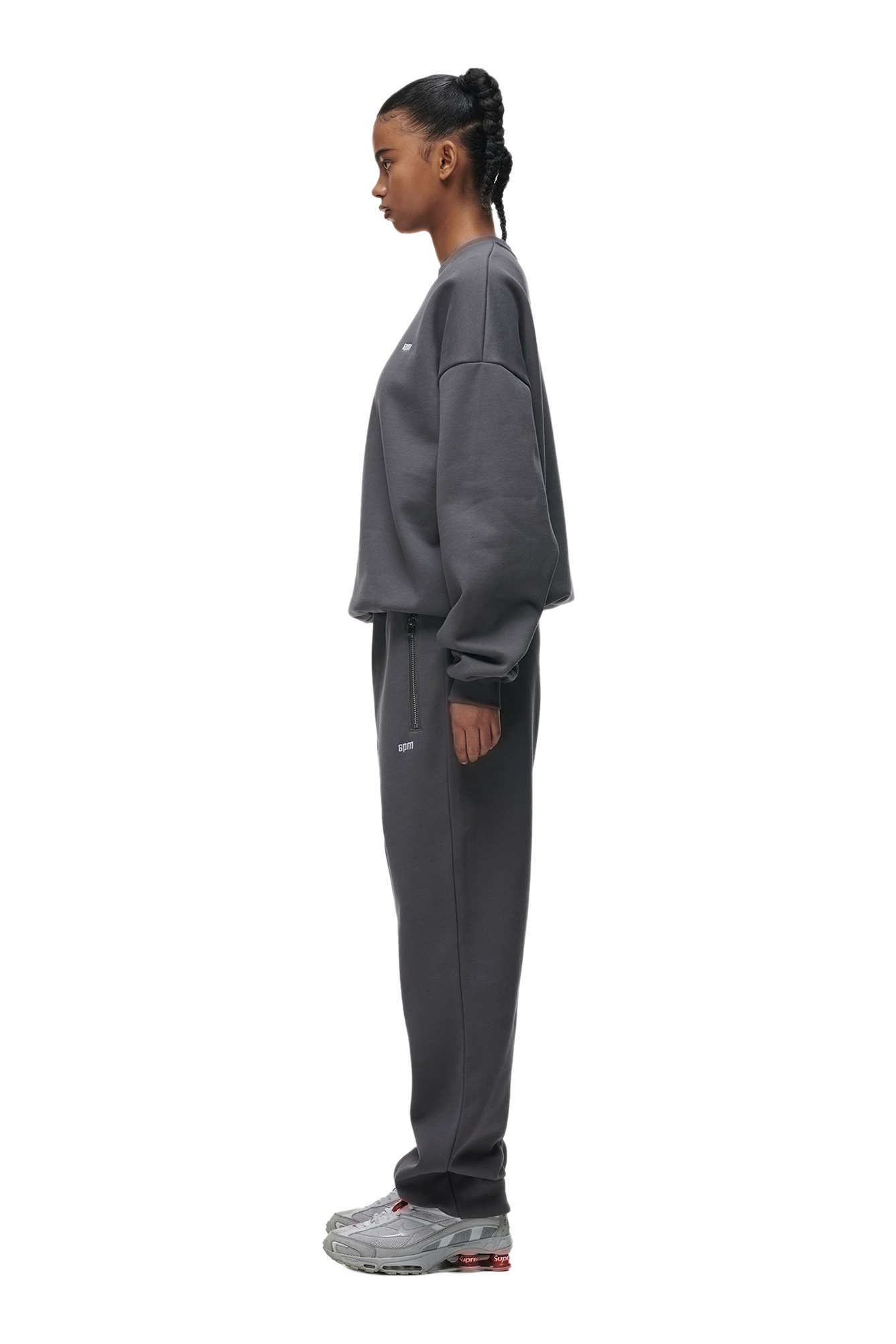 Someone stands in profile against a white background, wearing the 6PM CREWNECK ANTHRACITE sweatshirt with ribbed trims, matching sweatpants, and gray sneakers.