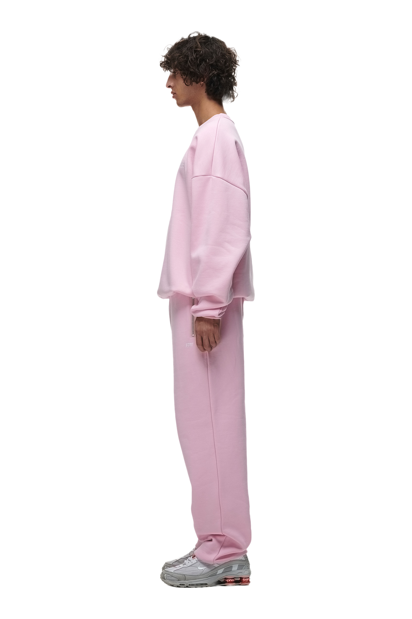 A person with curly hair stands in profile, wearing the 6PM CREWNECK BABY PINK sweatshirt and matching sweatpants with ribbed trims and gray sneakers, set against a white background.