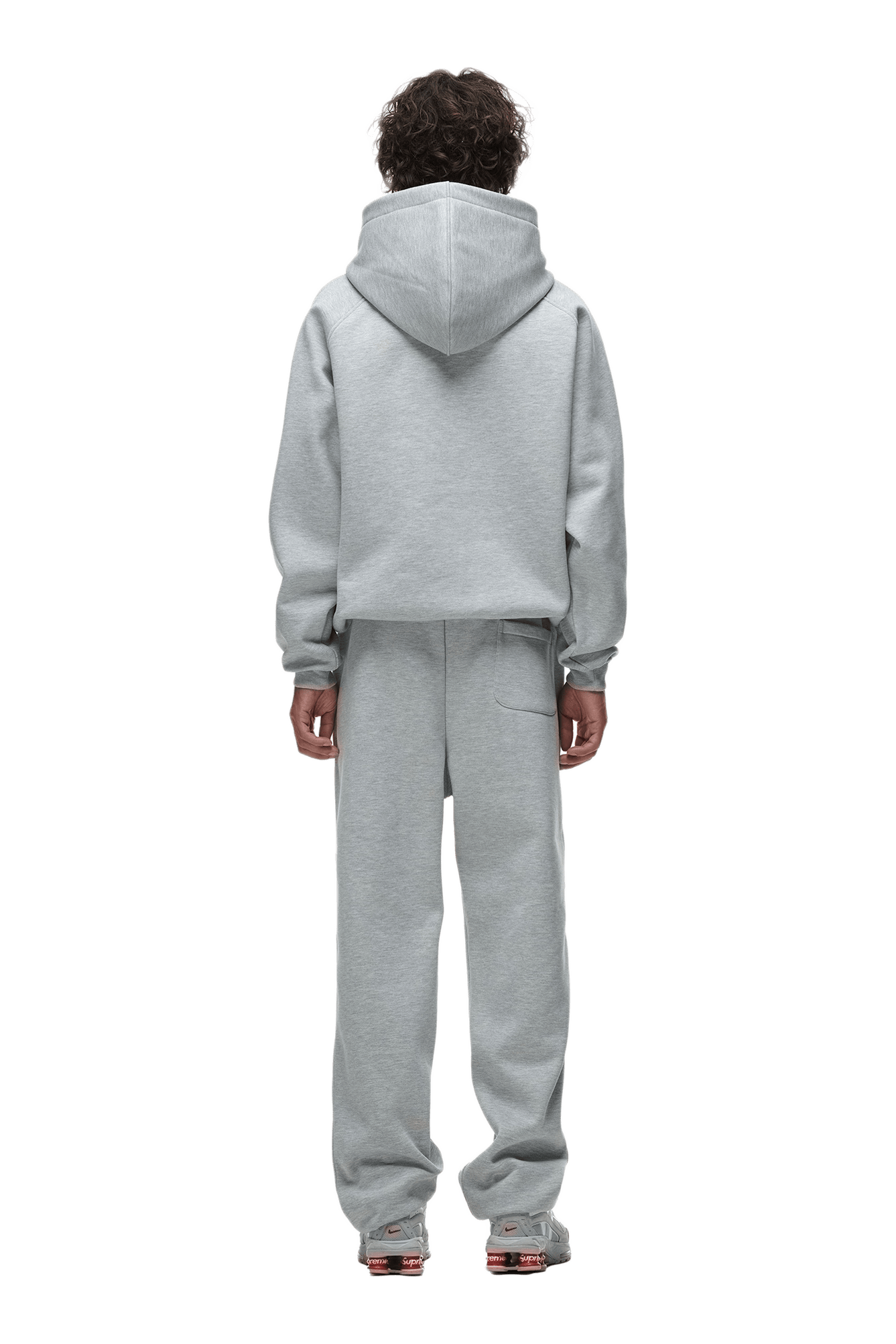 A person faces away from the camera in a 6PM HOODIE GREY MELANGE with raglan sleeves, hood up, kangaroo pocket, matching gray sweatpants, and sneakers. Transparent background—ideal for everyday use.