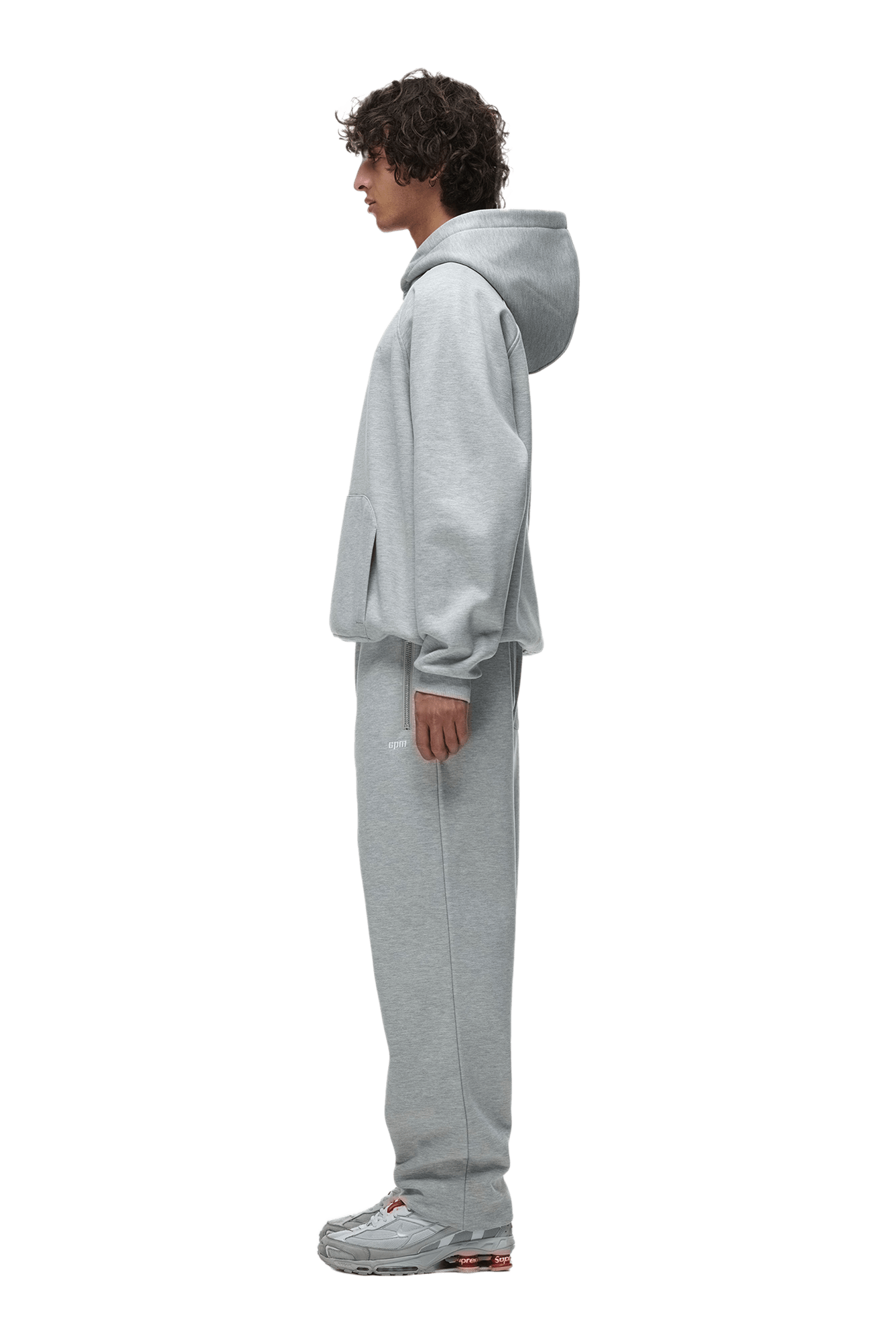 A person with curly hair stands in profile, wearing the 6PM HOODIE GREY MELANGE with Raglan sleeves and a kangaroo pocket, matching sweatpants, and gray sneakers—an ideal everyday look—against a transparent background.
