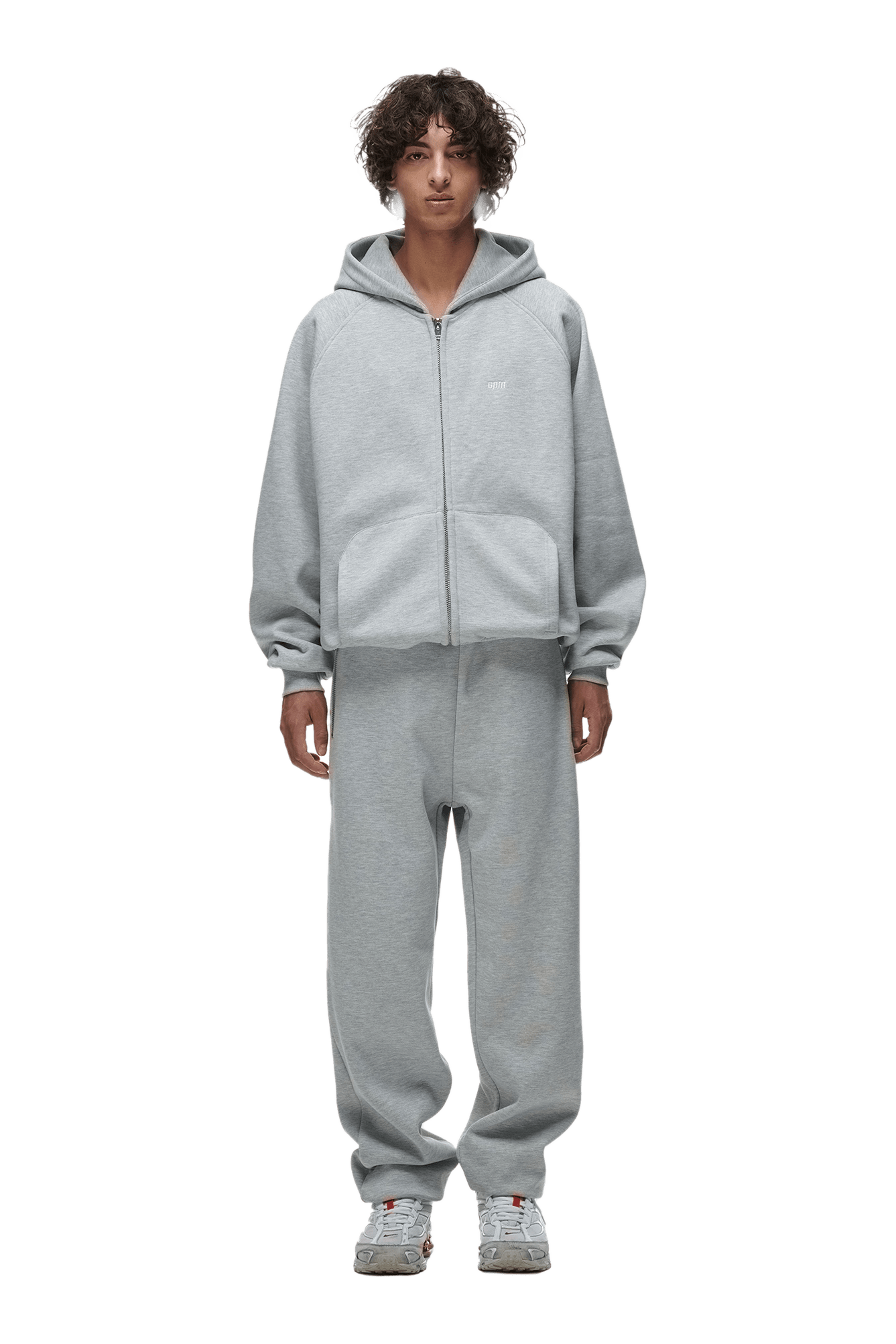 A person with curly hair wears the 6PM ZIP HOODIE GREY MELANGE, styled with matching sweatpants and gray sneakers, standing forward with hands by their sides.