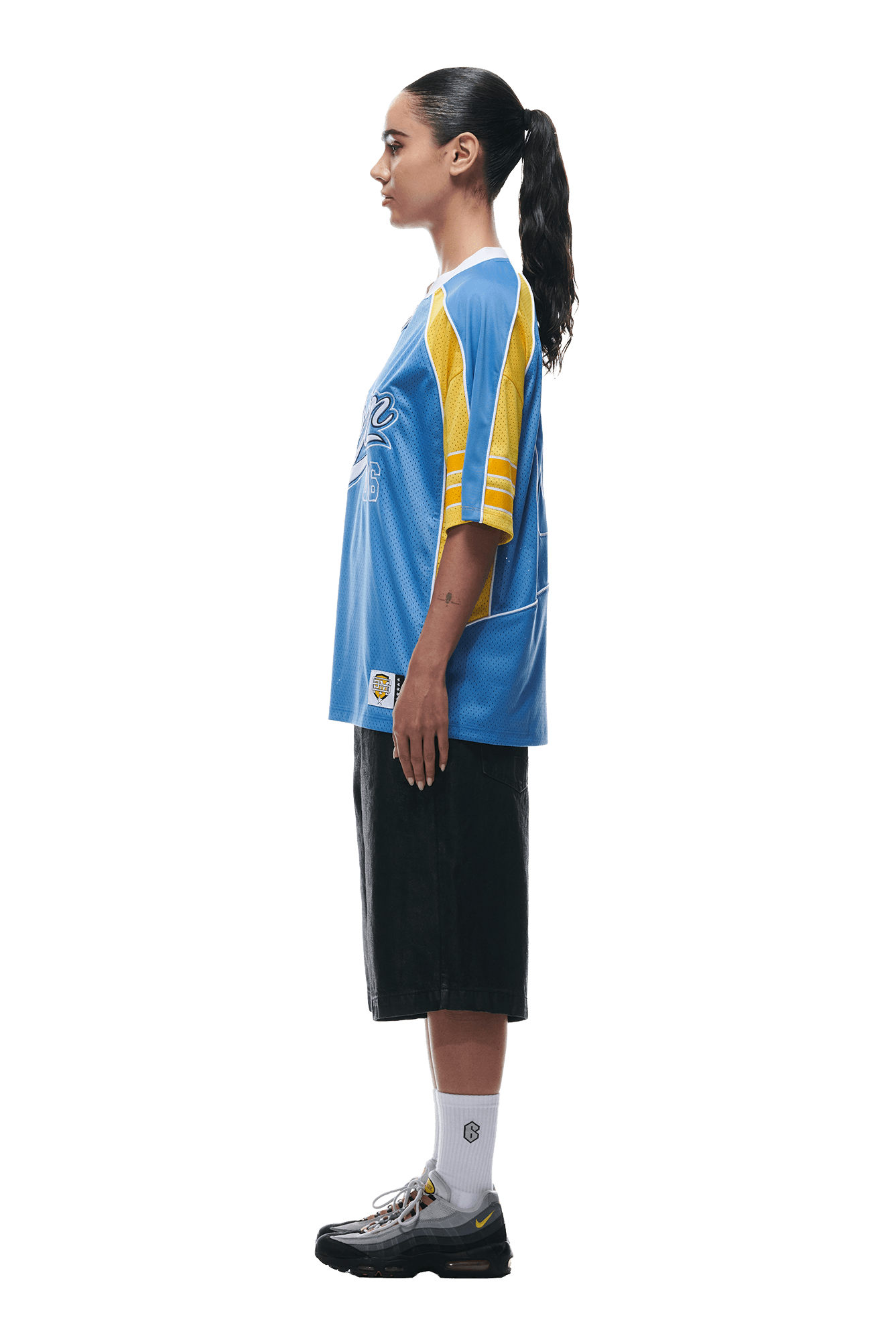 A person in profile wears the 6PM SUMMER FOOTBALL JERSEY BLUE, black knee-length shorts, white socks, and black sneakers; their long dark hair is tied back, with a plain white background.