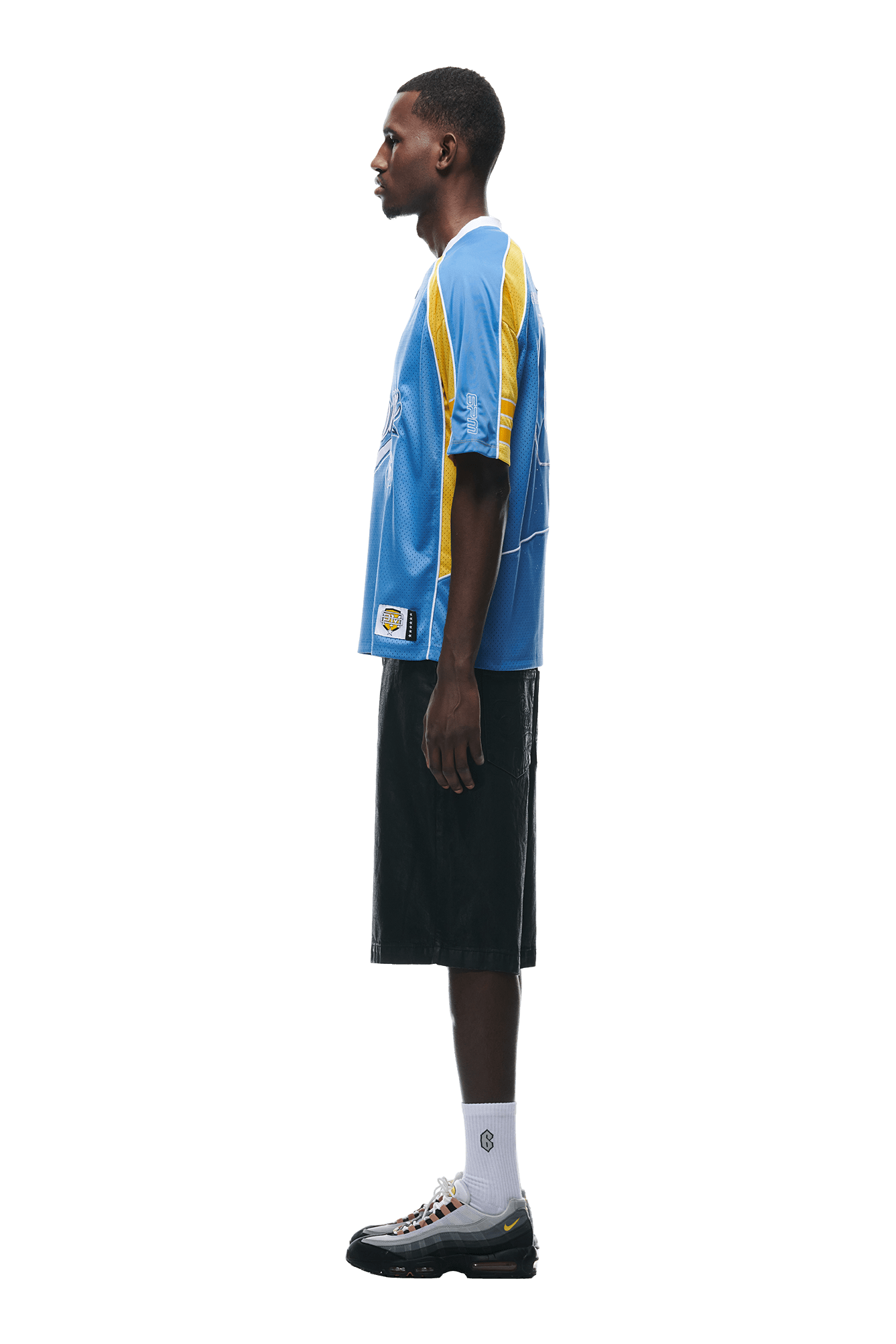 A man stands in profile against a white background, wearing the 6PM SUMMER FOOTBALL JERSEY BLUE with black knee-length shorts, white socks, and black sneakers.
