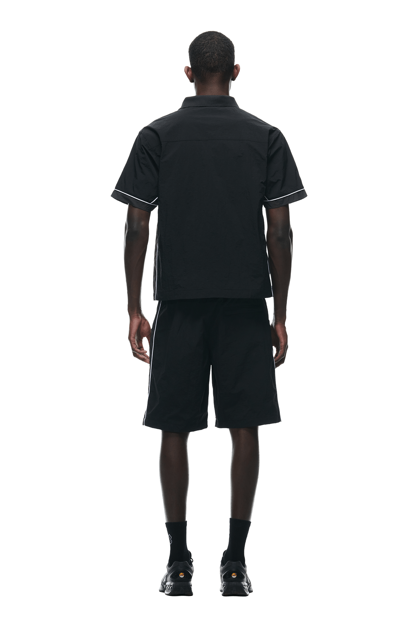 A man stands facing away from the camera wearing the 6PM WARM UP NYLON HALF ZIP BLACK, black shorts, socks, and sneakers against a plain white background.