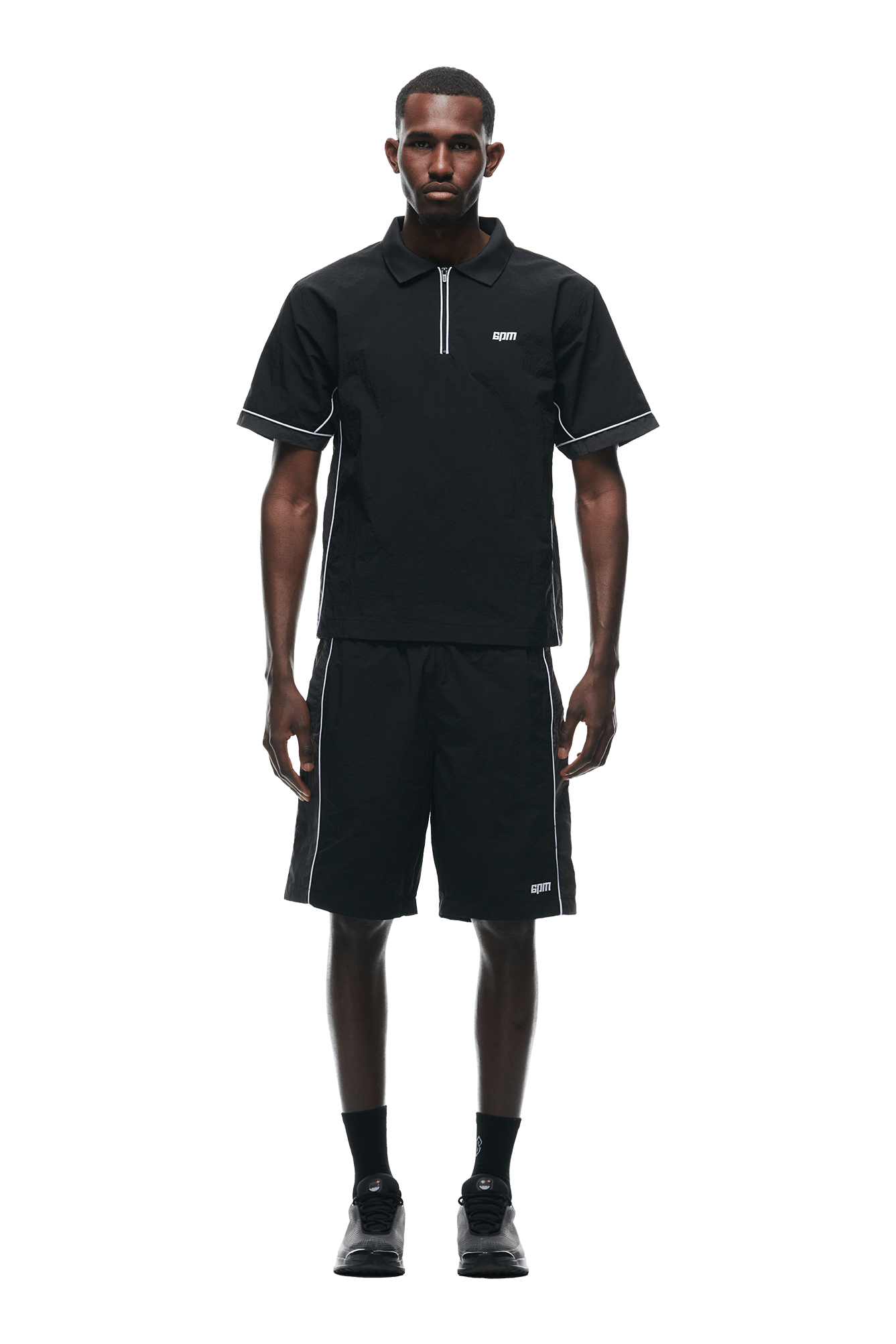 A man faces forward wearing the 6PM WARM UP NYLON HALF ZIP BLACK with matching black shorts featuring white piping, black socks, and black sneakers. The look is minimalist and sporty against a plain white background.