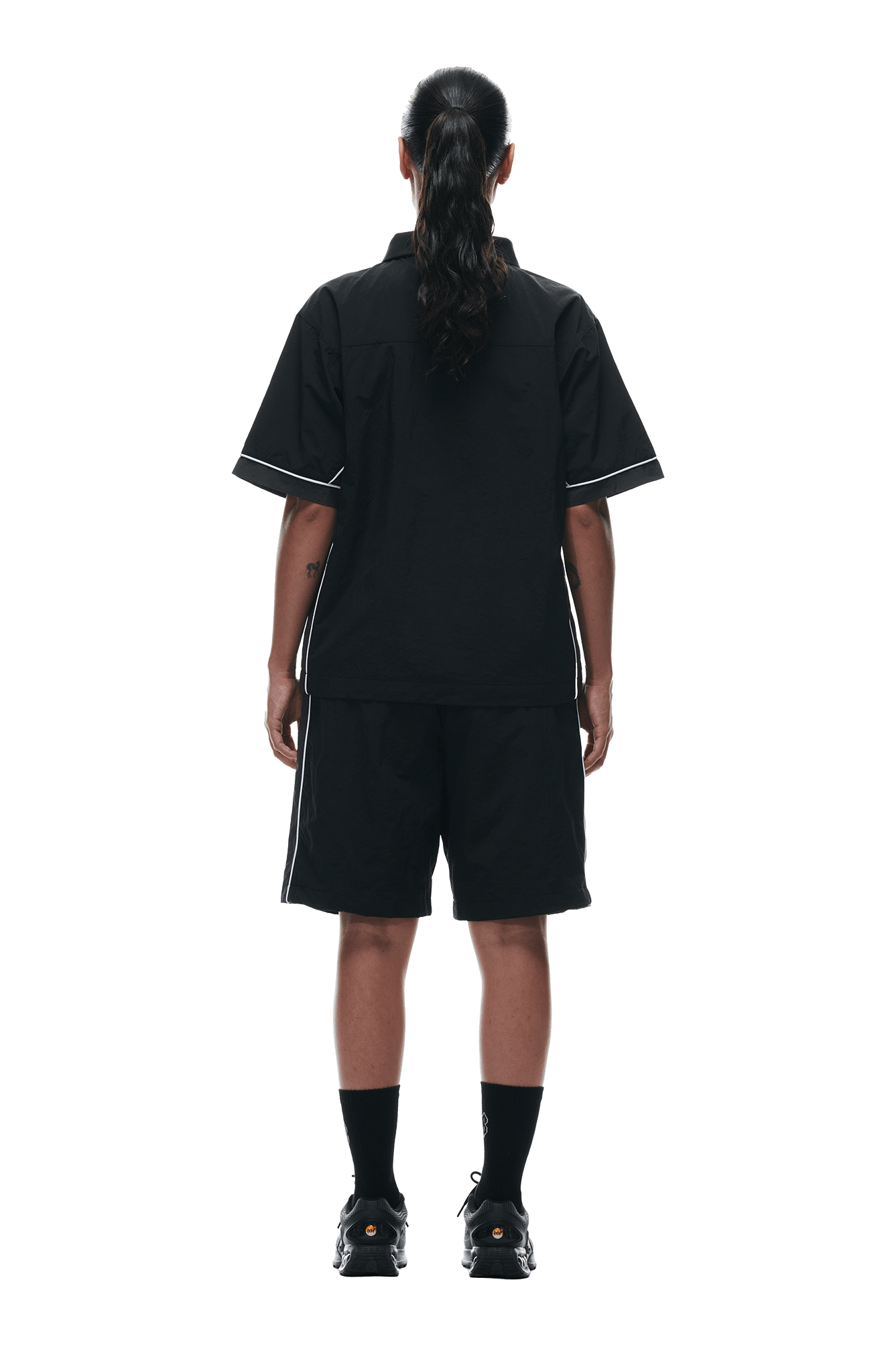 A person with long hair in a ponytail stands facing away, wearing the 6PM WARM UP NYLON HALF ZIP BLACK, black shorts, socks, and shoes, set against a plain white background.