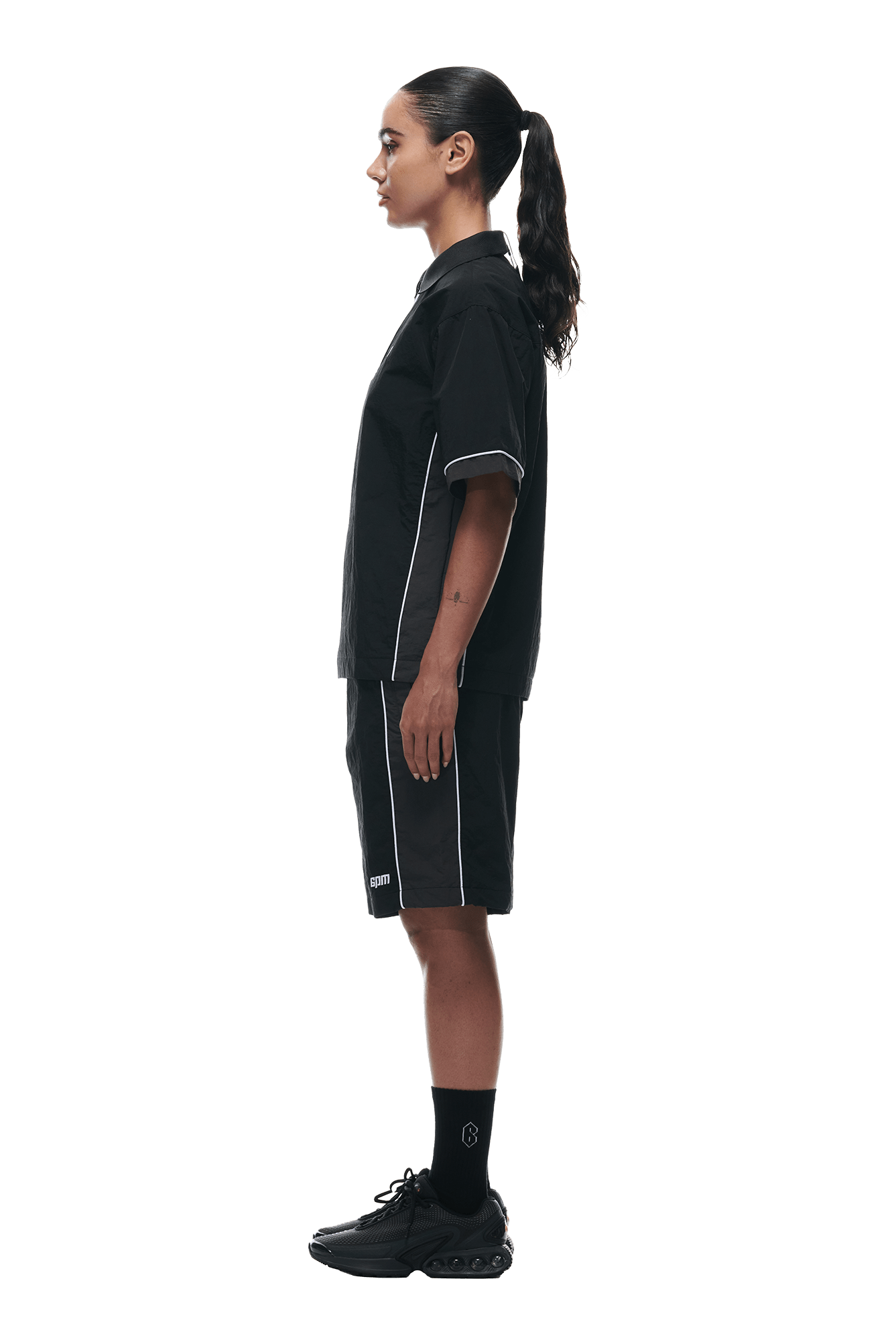 Wearing the 6PM WARM UP NYLON HALF ZIP BLACK, a person with a ponytail stands in profile against a white background, styled with matching black shorts, black socks, and black sneakers.