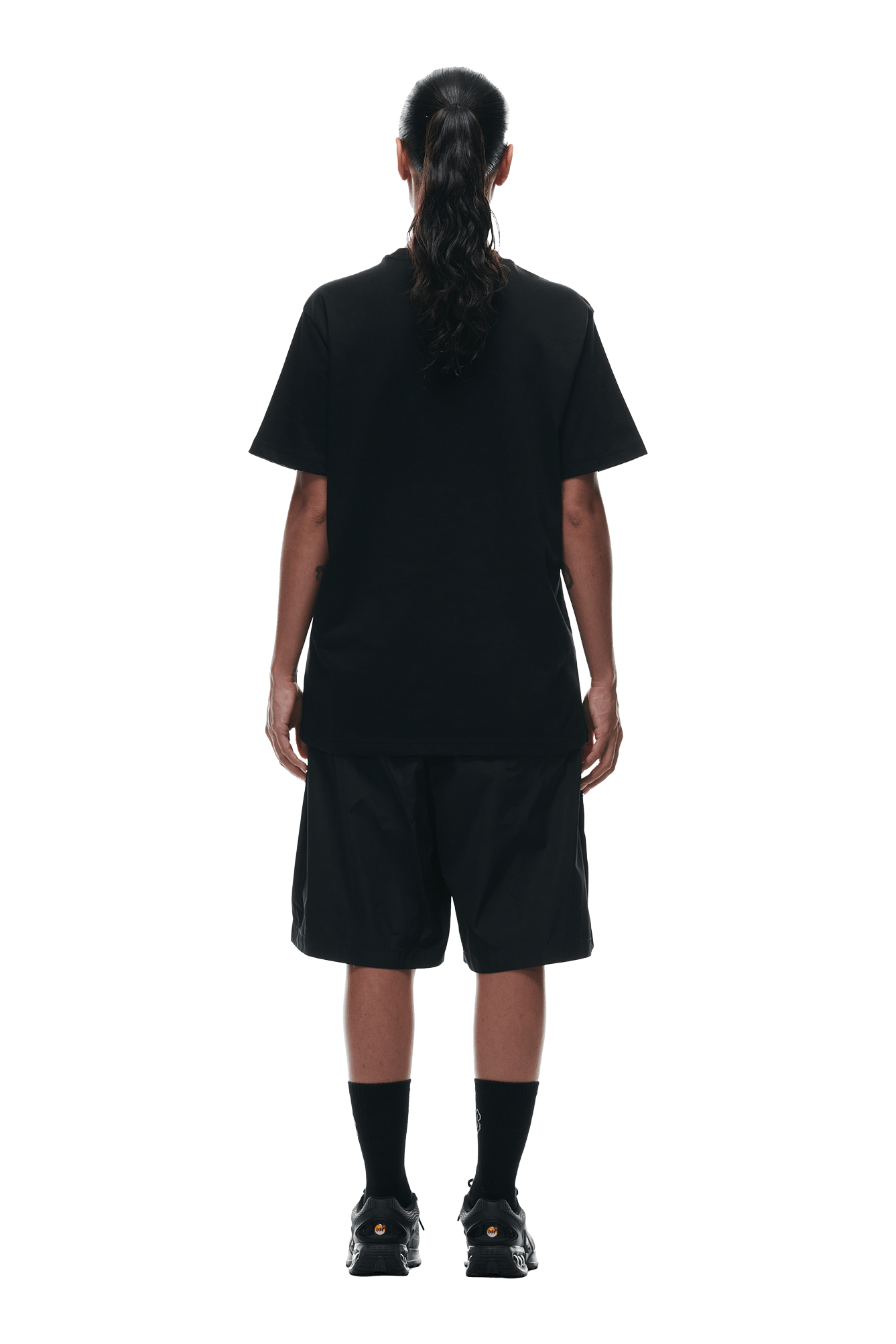 Person with long dark hair in a ponytail wears the 6PM BUBBLE LOGO T-SHIRT BLACK, black shorts, socks, and sneakers, standing with their back to the camera against a plain white background.