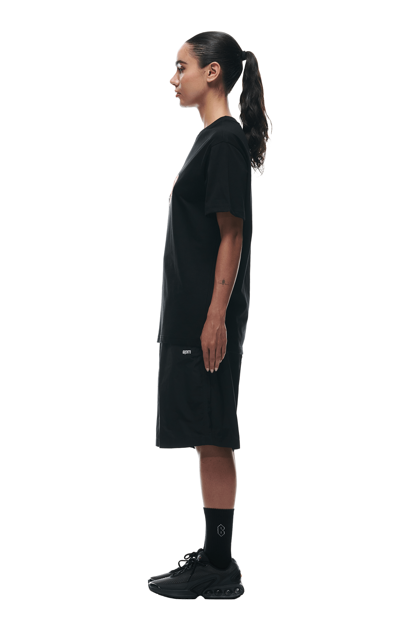 A person with long dark hair in a ponytail stands in profile against a white background, wearing the 6PM BUBBLE LOGO T-SHIRT BLACK, paired with black shorts, socks, and sneakers.