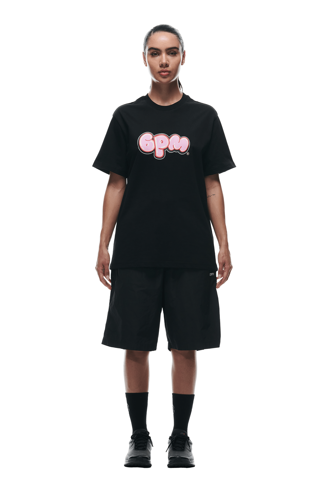 A person stands facing forward in a 6PM BUBBLE LOGO T-SHIRT BLACK, black knee-length shorts, black socks, and black sneakers, against a plain white background.