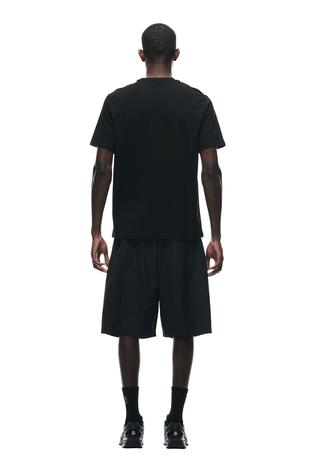 A man stands facing away, wearing a 6PM BUBBLE LOGO T-SHIRT BLACK, loose black shorts, black socks, and black sneakers, set against a white background.