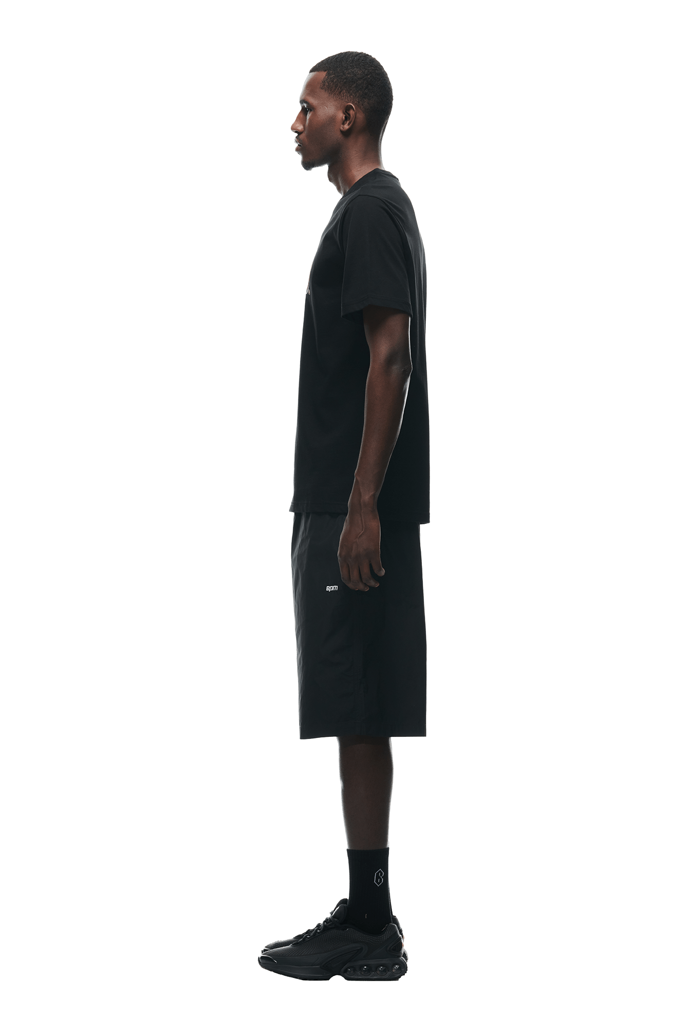 A man in profile faces left, wearing the 6PM BUBBLE LOGO T-SHIRT BLACK with black shorts, socks, and sneakers against a plain white background.
