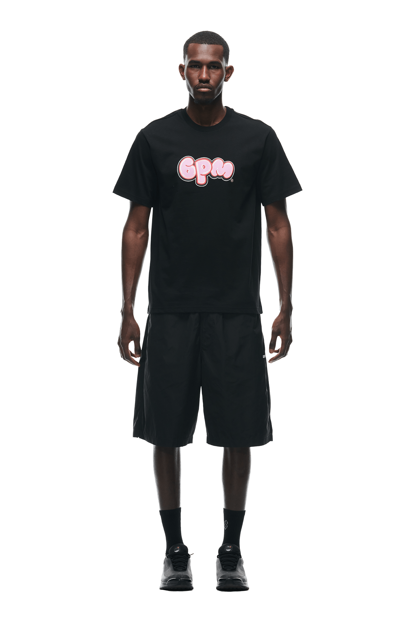 A man stands facing forward in a 6PM BUBBLE LOGO T-SHIRT BLACK, black knee-length shorts, black socks, and black sneakers against a plain white background.