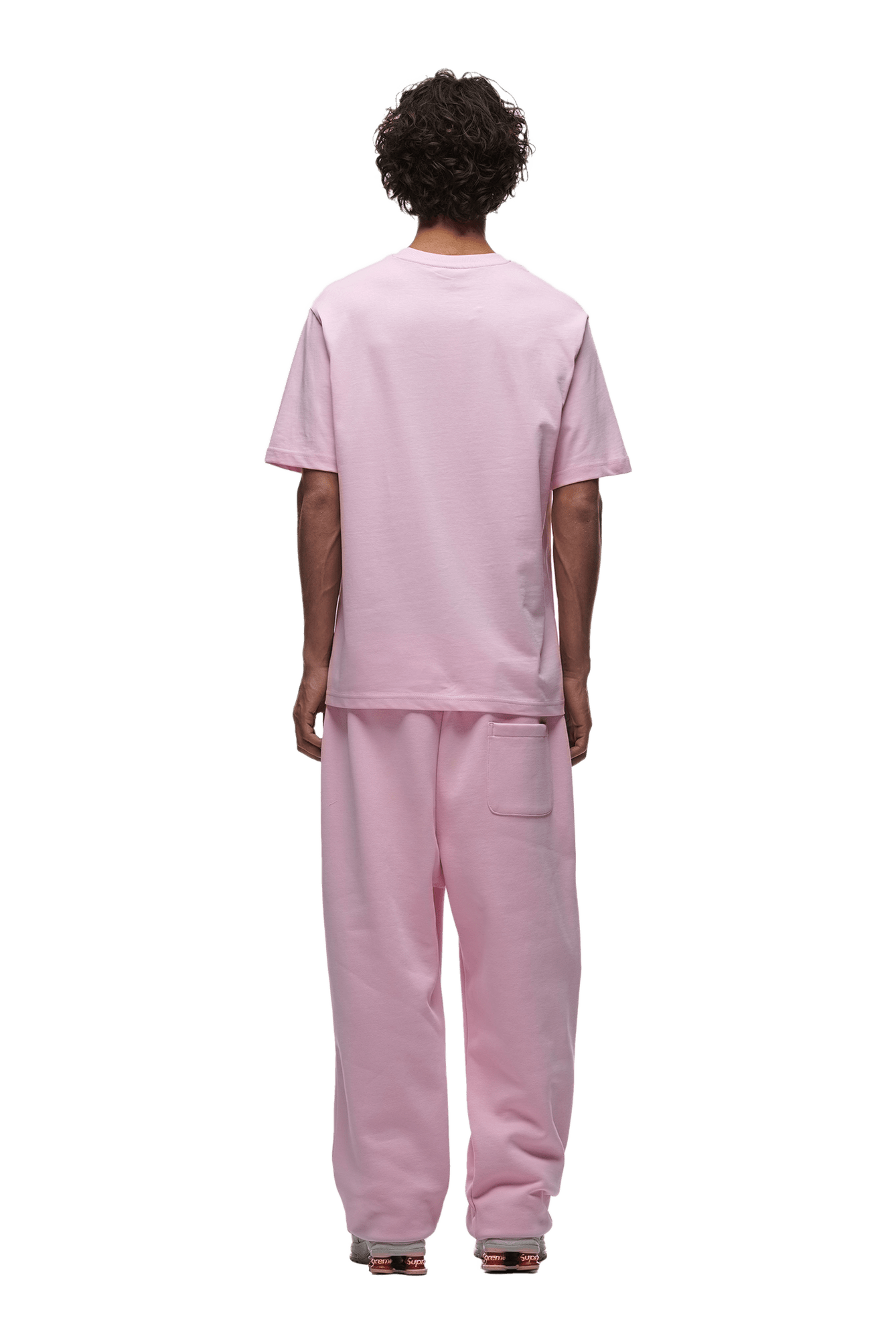 A person with curly hair faces away, wearing the 6PM T-SHIRT BABY PINK and matching pink sweatpants with white shoes, all shown on a transparent background.