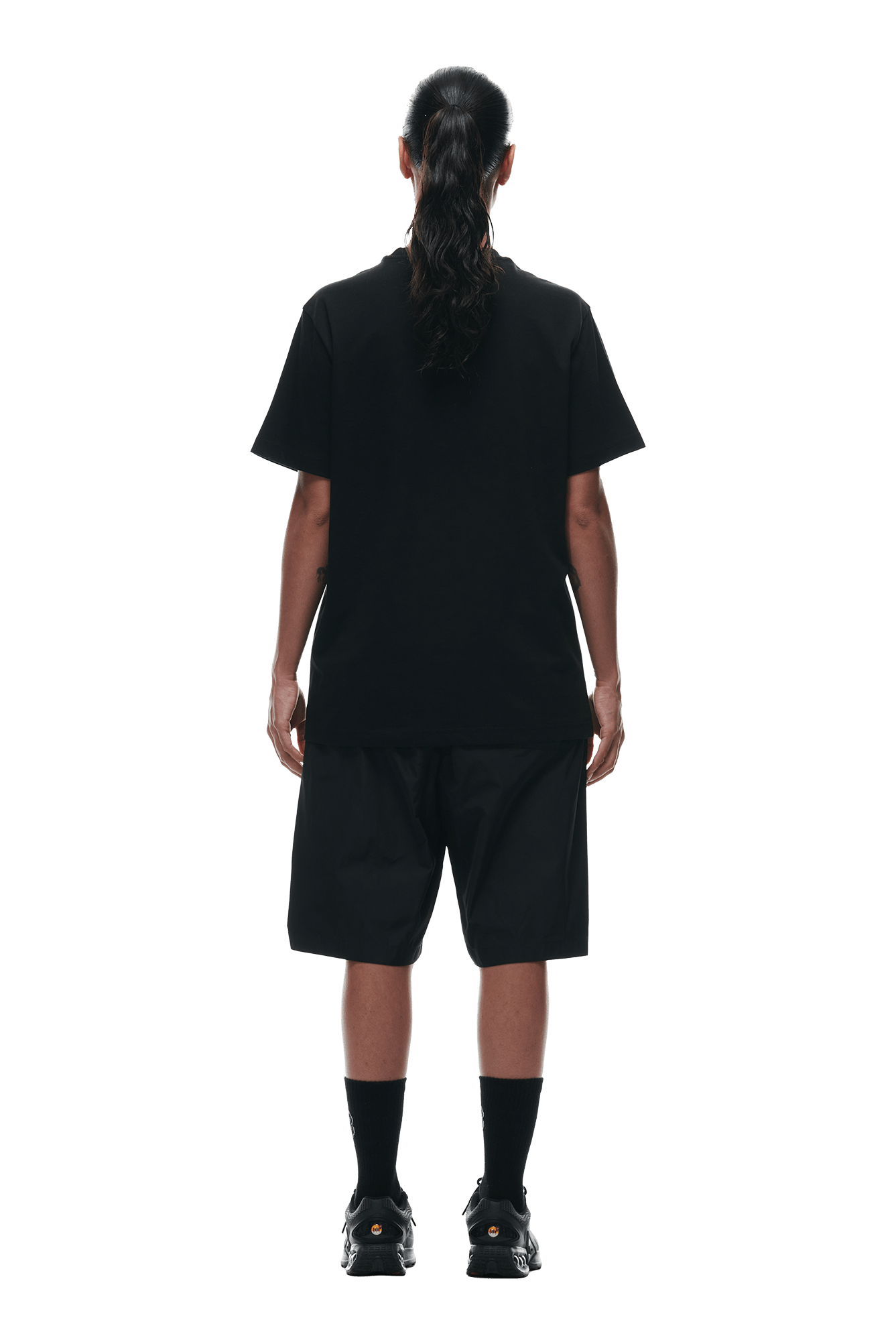 Person with long dark hair in a ponytail, wearing the 6PM GEMS T-SHIRT BLACK, loose black shorts, black socks, and black sneakers, stands with their back to the camera against a white background.