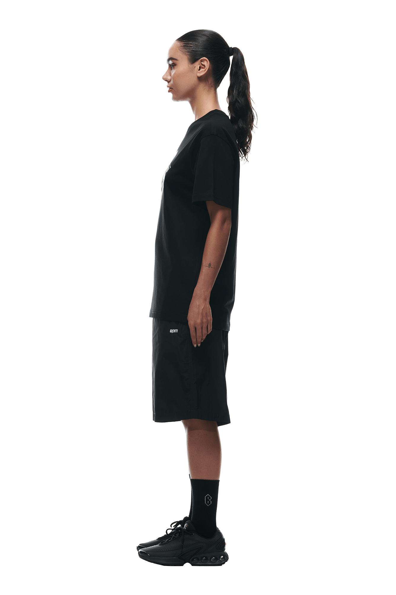 A person with long dark hair in a ponytail stands in profile against a white background, wearing the 6PM GEMS T-SHIRT BLACK, black shorts, black socks, and black sneakers.