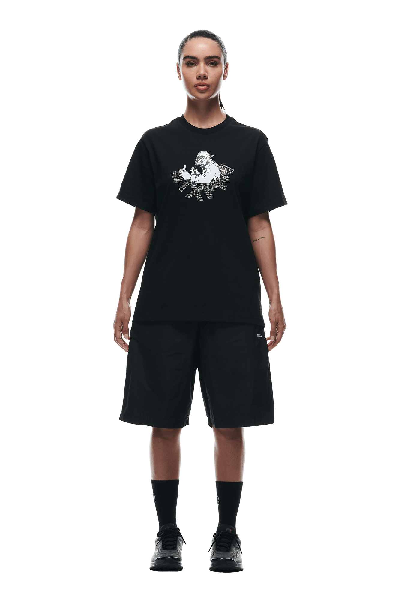 A person faces forward in a 6PM GEMS T-SHIRT BLACK, paired with loose black shorts, black socks, and black sneakers. Their hair is tied back and the background is plain white.
