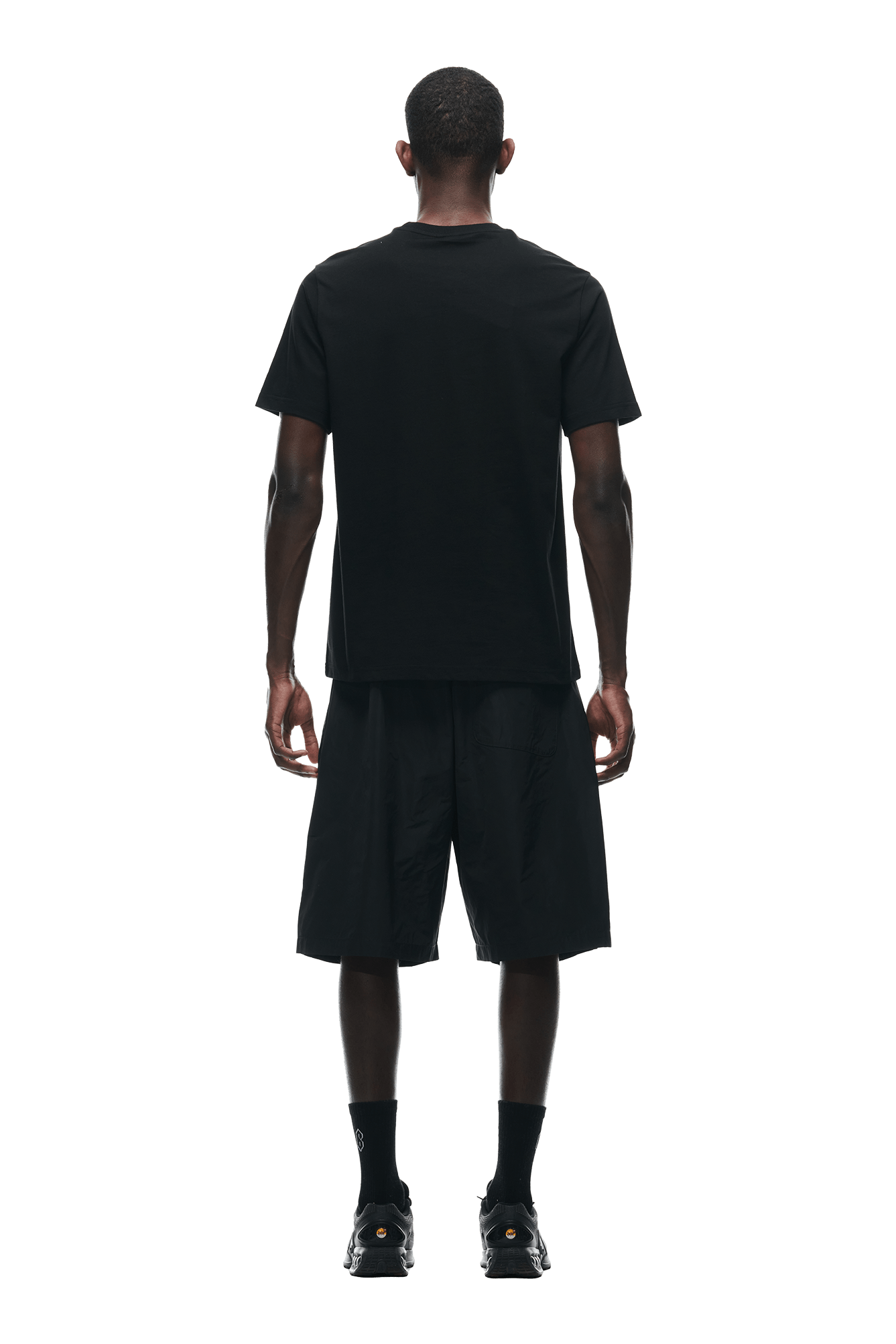 A man faces away from the camera wearing the 6PM GEMS T-SHIRT BLACK, paired with black shorts, socks, and sneakers, set against a plain white background.