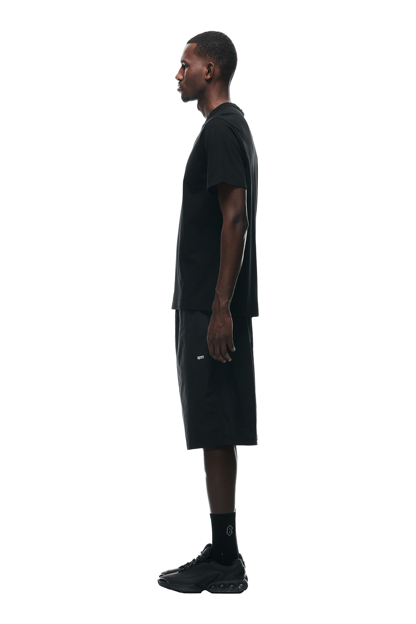 A man stands in profile against a white background, wearing the 6PM GEMS T-SHIRT BLACK, paired with black shorts, socks, and sneakers.