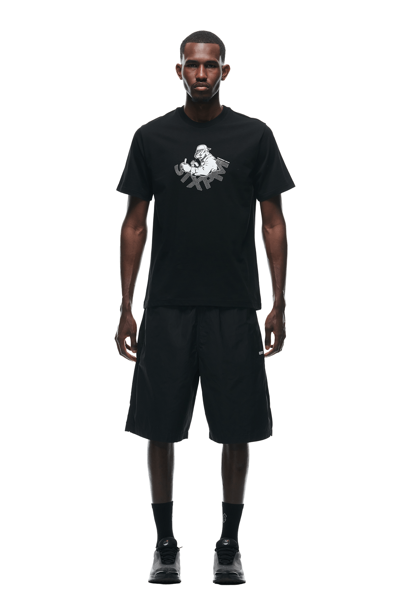 A man faces forward against a white background, wearing the 6PM GEMS T-SHIRT BLACK, loose black shorts, black socks, and black sneakers.