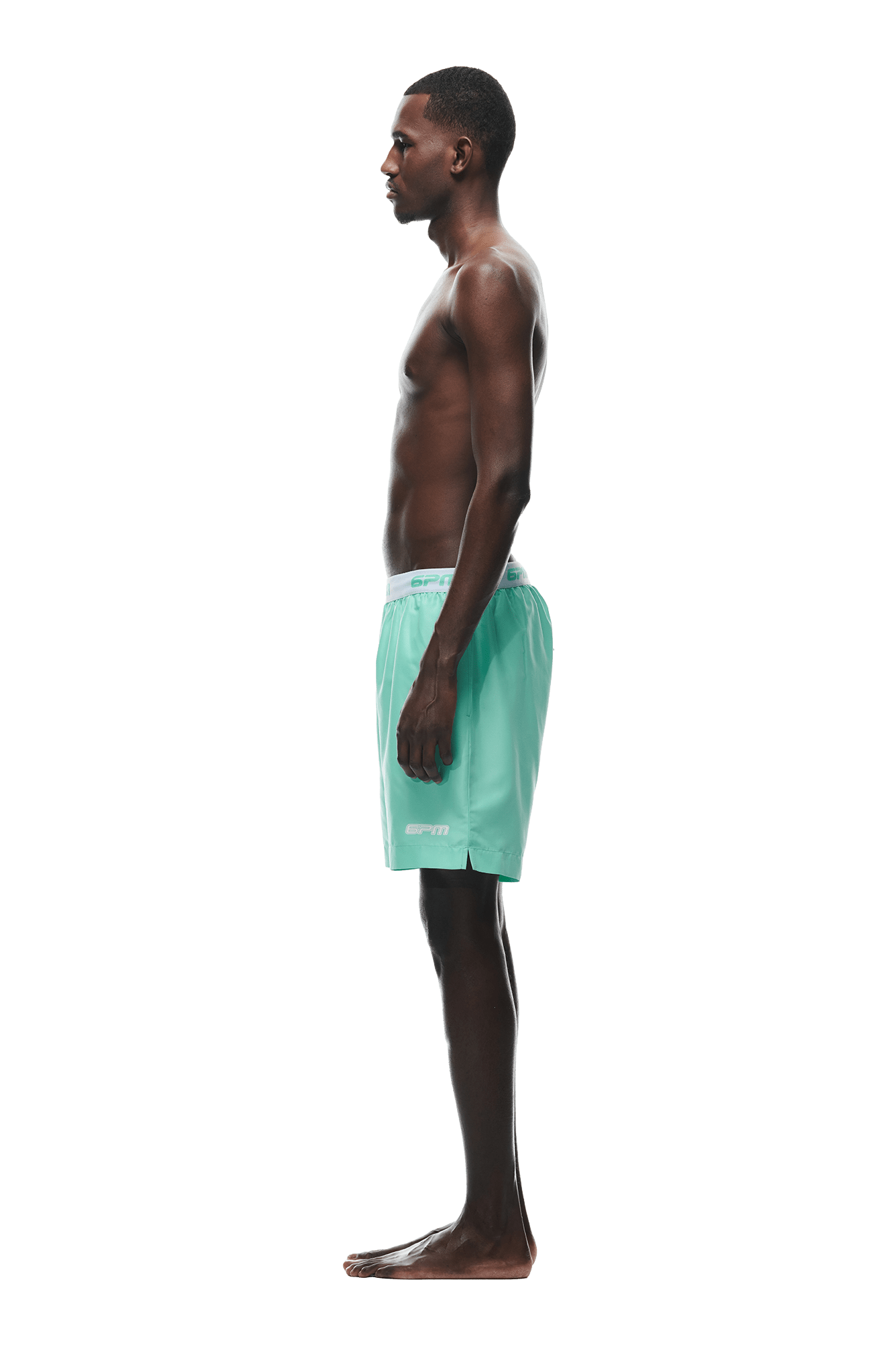 A man stands in profile against a white background, barefoot and shirtless, wearing 6PM BOXER SWIM SHORTS in Light Moss. His arms are relaxed at his sides.