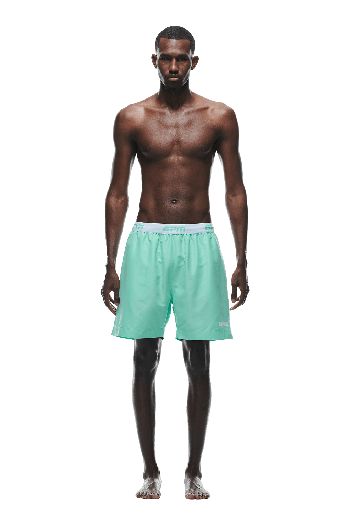 A man stands barefoot against a white background, wearing 6PM BOXER SWIM SHORTS LIGHT MOSS with a gray waistband and white logo. He is shirtless, facing forward with a neutral expression.