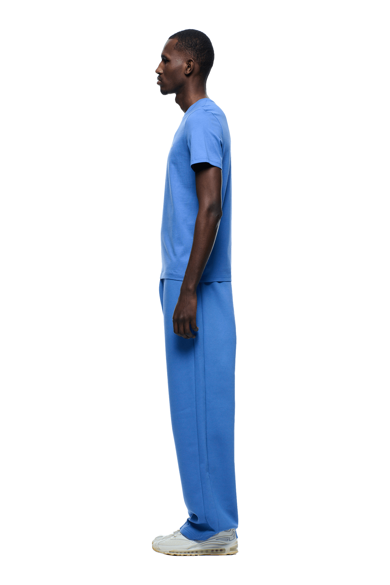 A man stands in profile facing left, wearing the 6PM SLIM FIT T-SHIRT PERSIAN BLUE with matching blue sweatpants and white sneakers, set against a plain background.